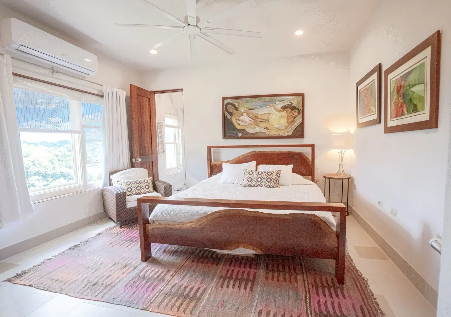 A bright bedroom with a large bed covered in a white quilt, flanked by pillows, and a wooden headboard. A window with white curtains allows natural light. There is a patterned rug on the floor, a small side table, and framed artwork on the walls. A flat-screen TV is mounted on the wall opposite the bed.