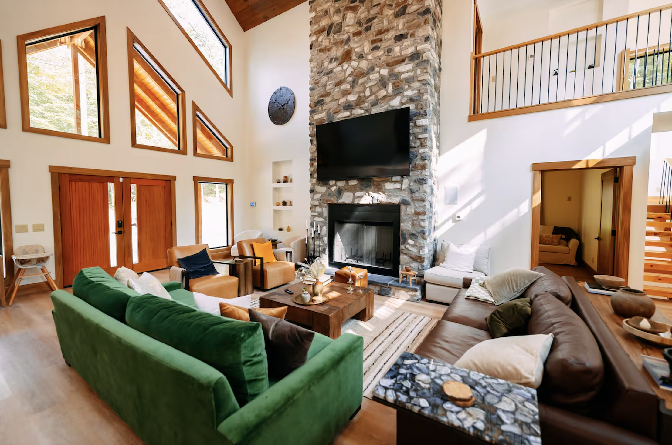 A spacious living room with high ceilings, large triangular windows, and a stone fireplace with a flat screen TV mounted above. The room features a mix of colorful and neutral sofas, a wooden coffee table, and warm wood accents.