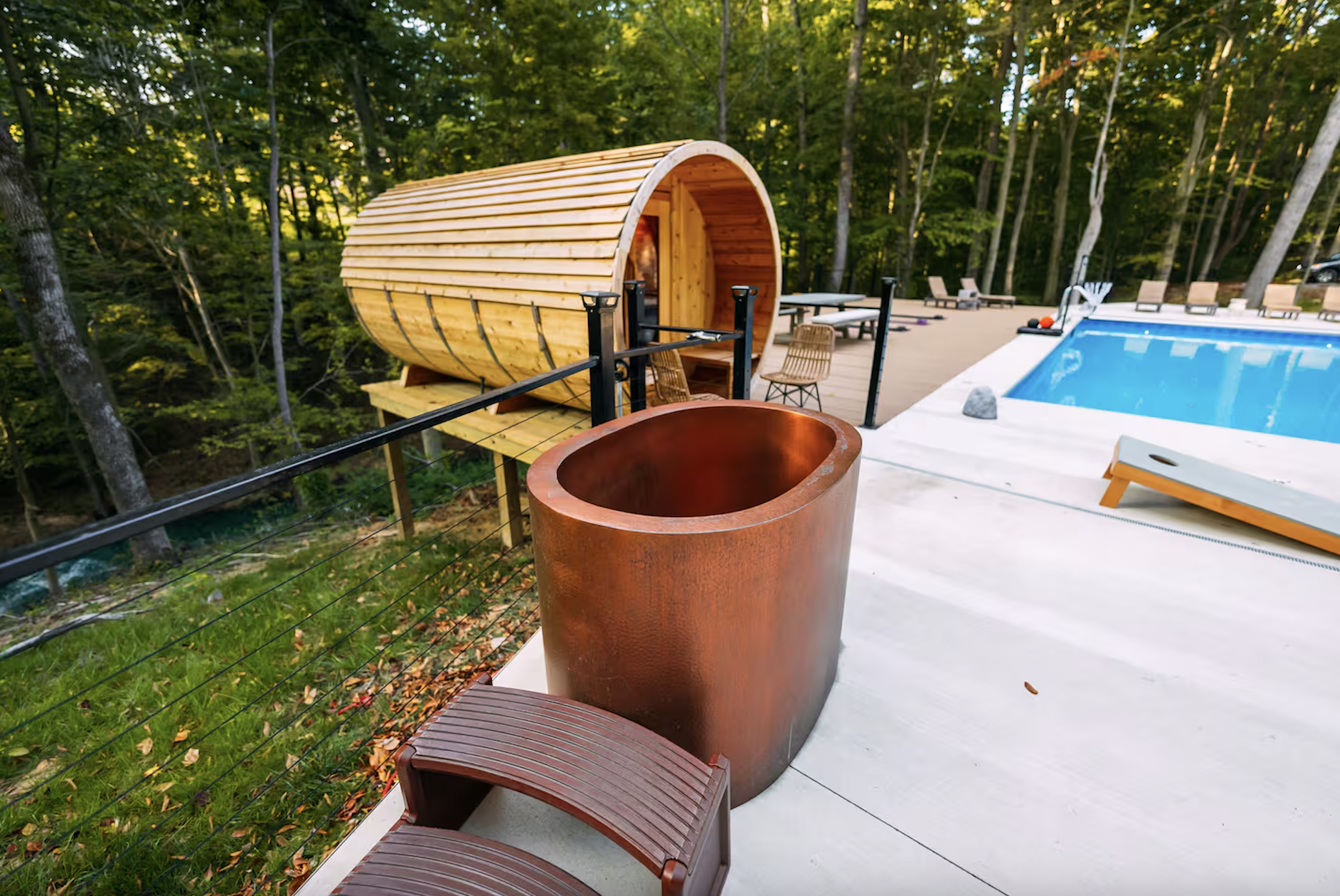 A modern backyard with a swimming pool, a wooden sauna, and a rust-colored outdoor bathtub amid a wooded area.