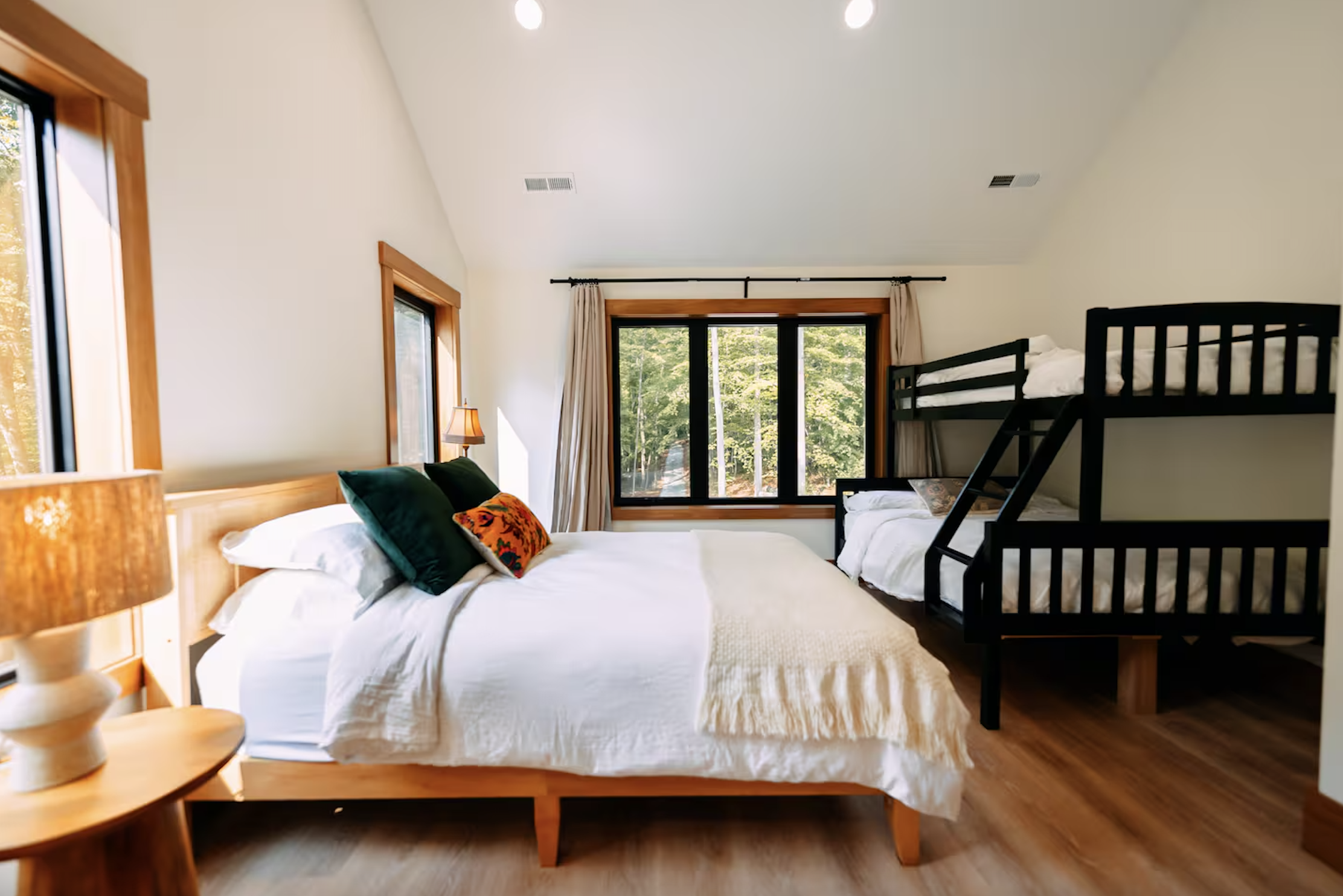 Bright bedroom with a large bed, pillows, a small wooden side table, a lamp, and a bunk bed near a large window with curtains showing a view of trees outside.