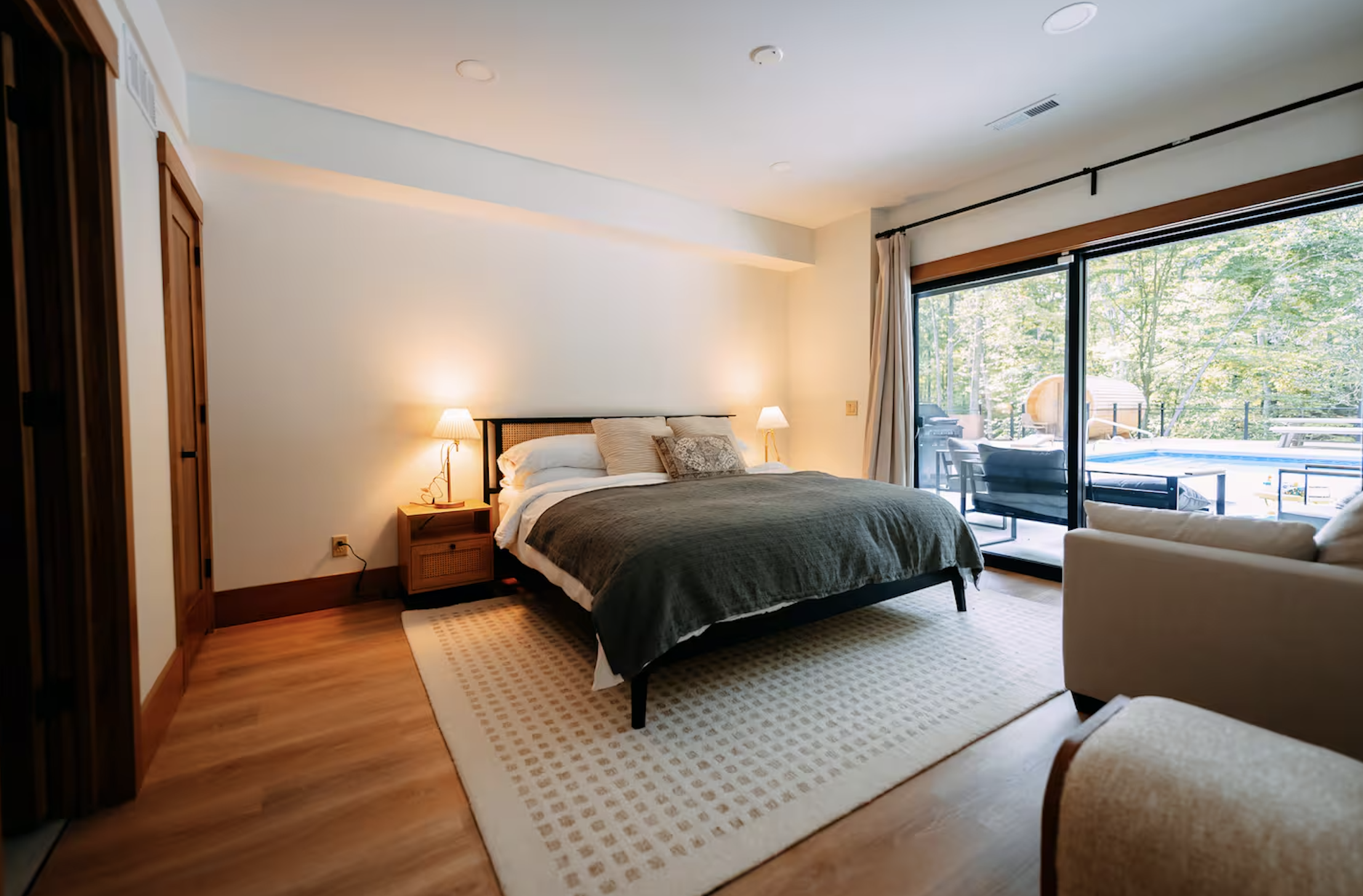 Bedroom with large bed, nightstands, lamps, sliding glass door leading to patio with outdoor furniture and greenery outside.