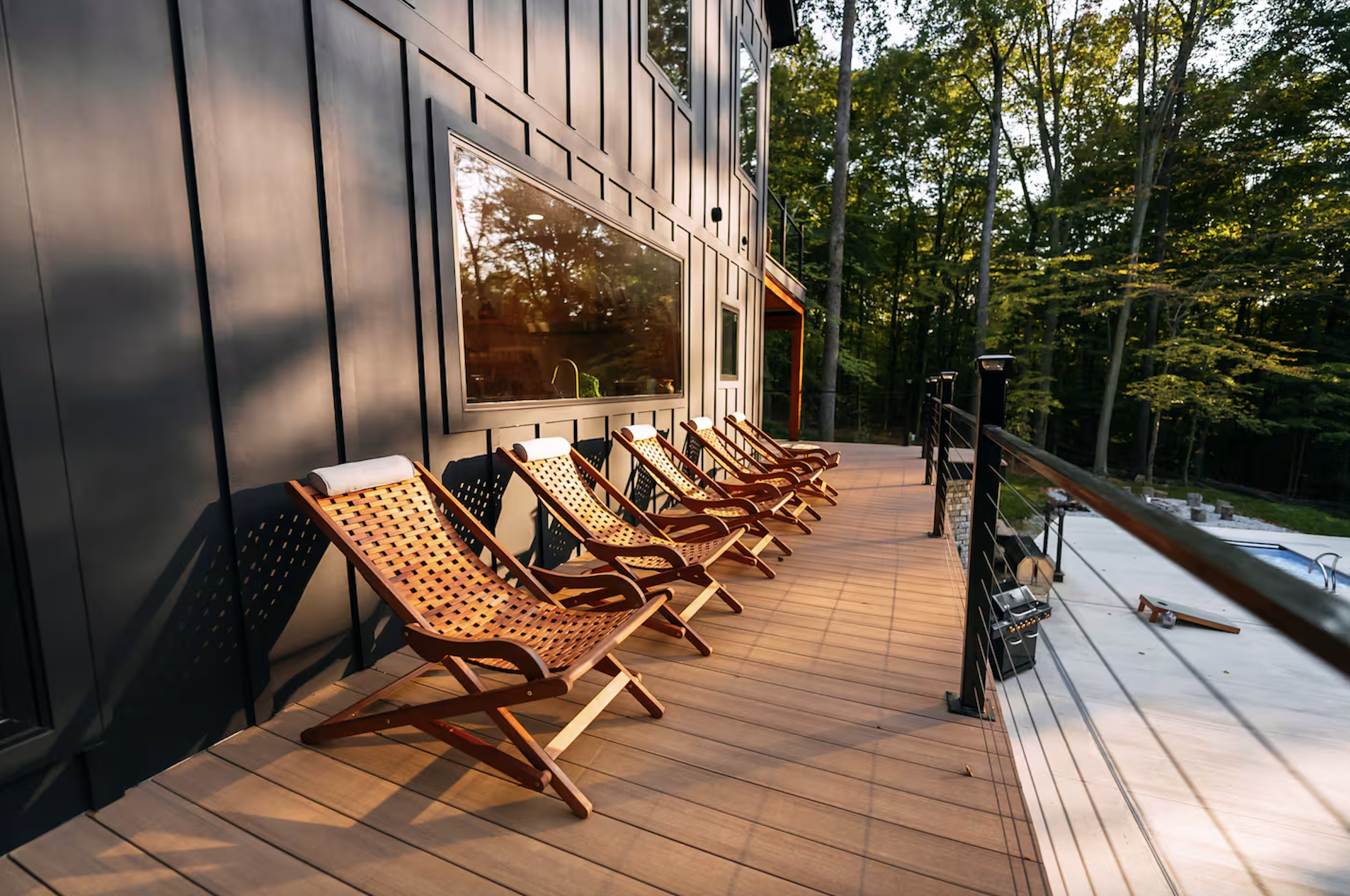 Outdoor wooden deck with five wooden lounge chairs with towels on top, overlooking trees at sunset or sunrise.