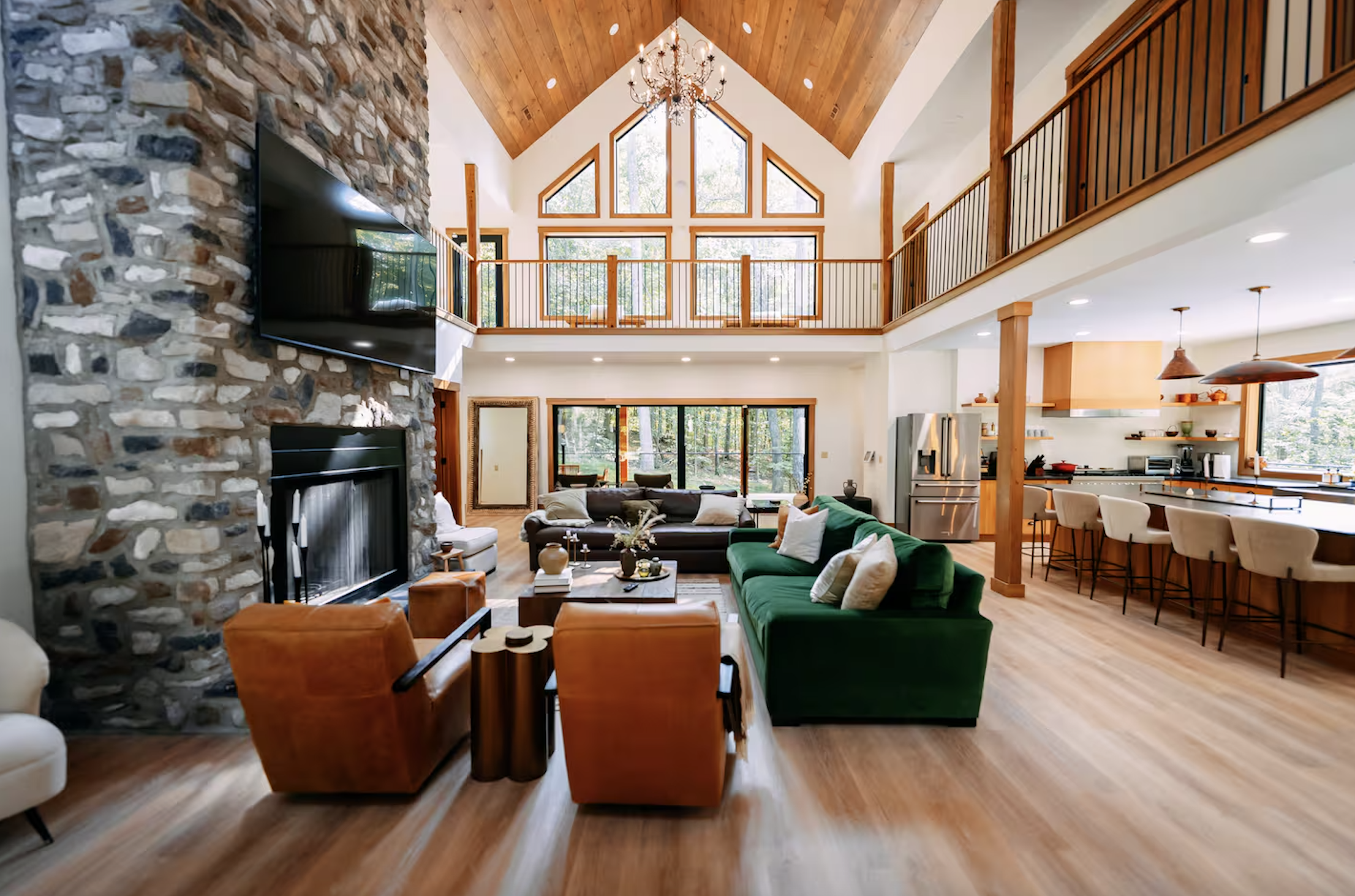 Spacious living room with high vaulted ceiling, large windows, stone fireplace, and modern furniture including green and brown sofas, with a view into the open kitchen area.