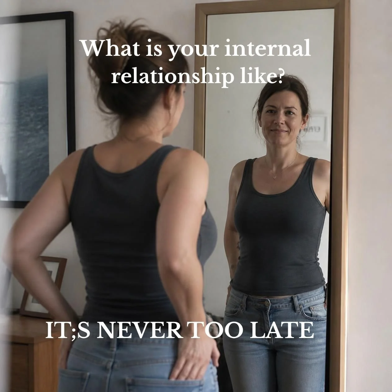 A Woman with dark hair standing in front of a full length mirror looking at herself.  She is wearing a dark sleeveless top and jeans with her hands on her hips.