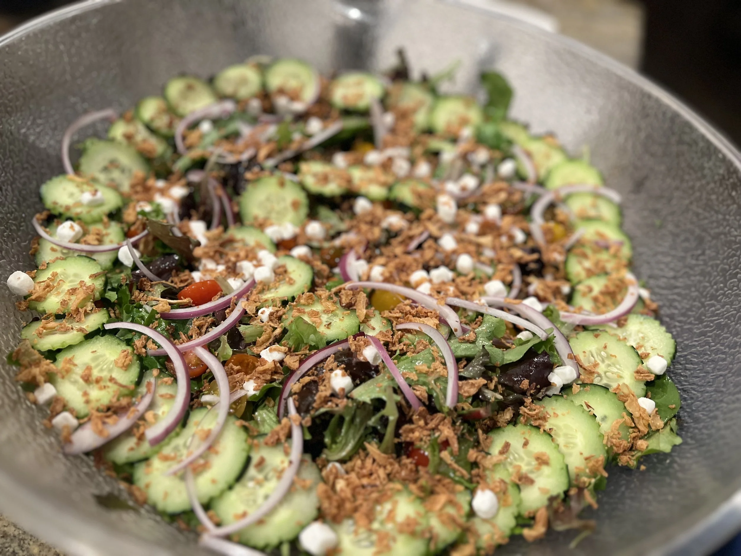 Fresh mixed salad with sliced cucumbers, red onions, cherry tomatoes, chopped lettuce, and cheese crumbles, topped with crispy fried onion pieces in a glass bowl.