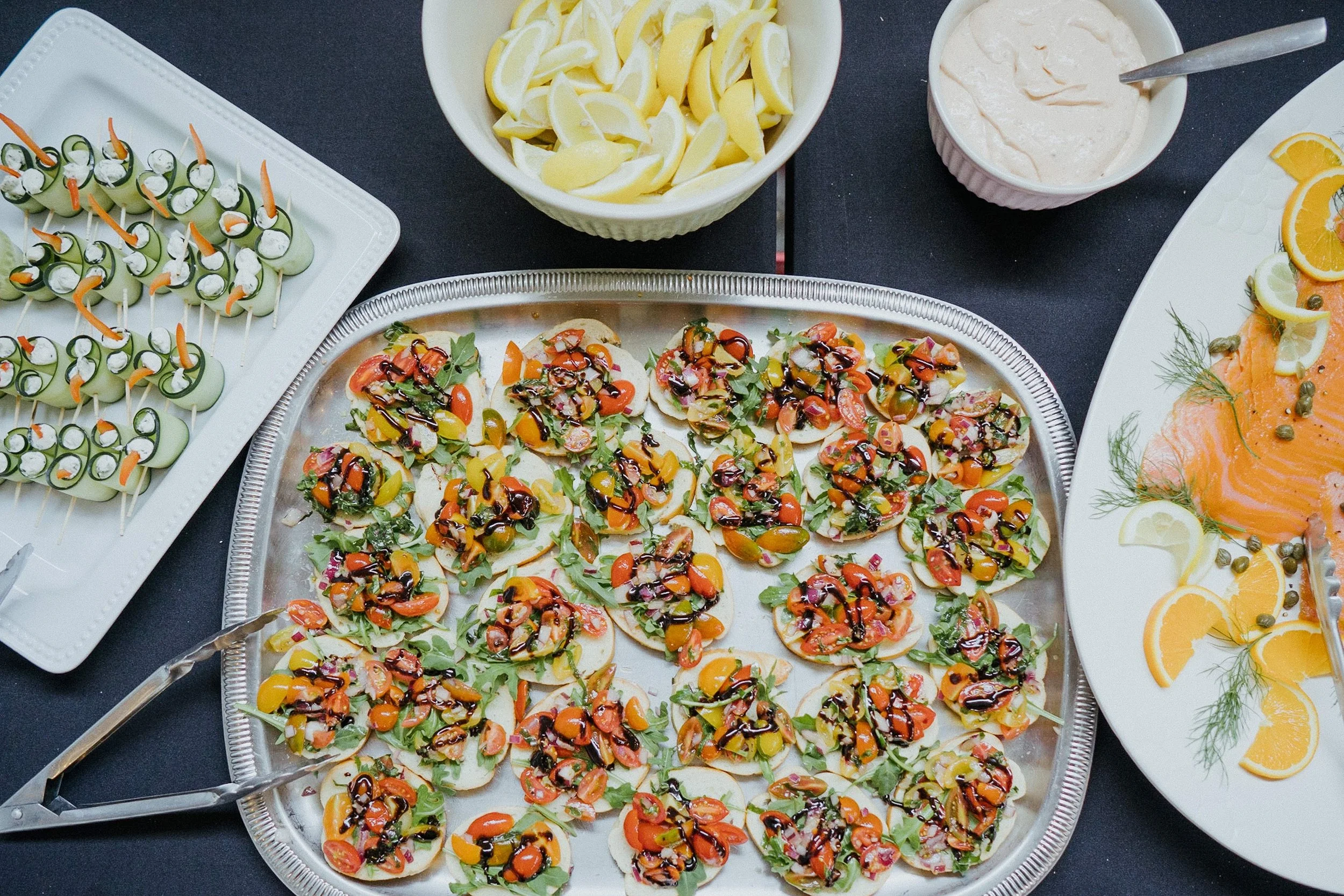 A variety of appetizers including small sandwiches with toppings, cucumber and cream cheese skewers, lemon wedges, smoked salmon with lemon slices and capers, and a bowl of creamy dip.