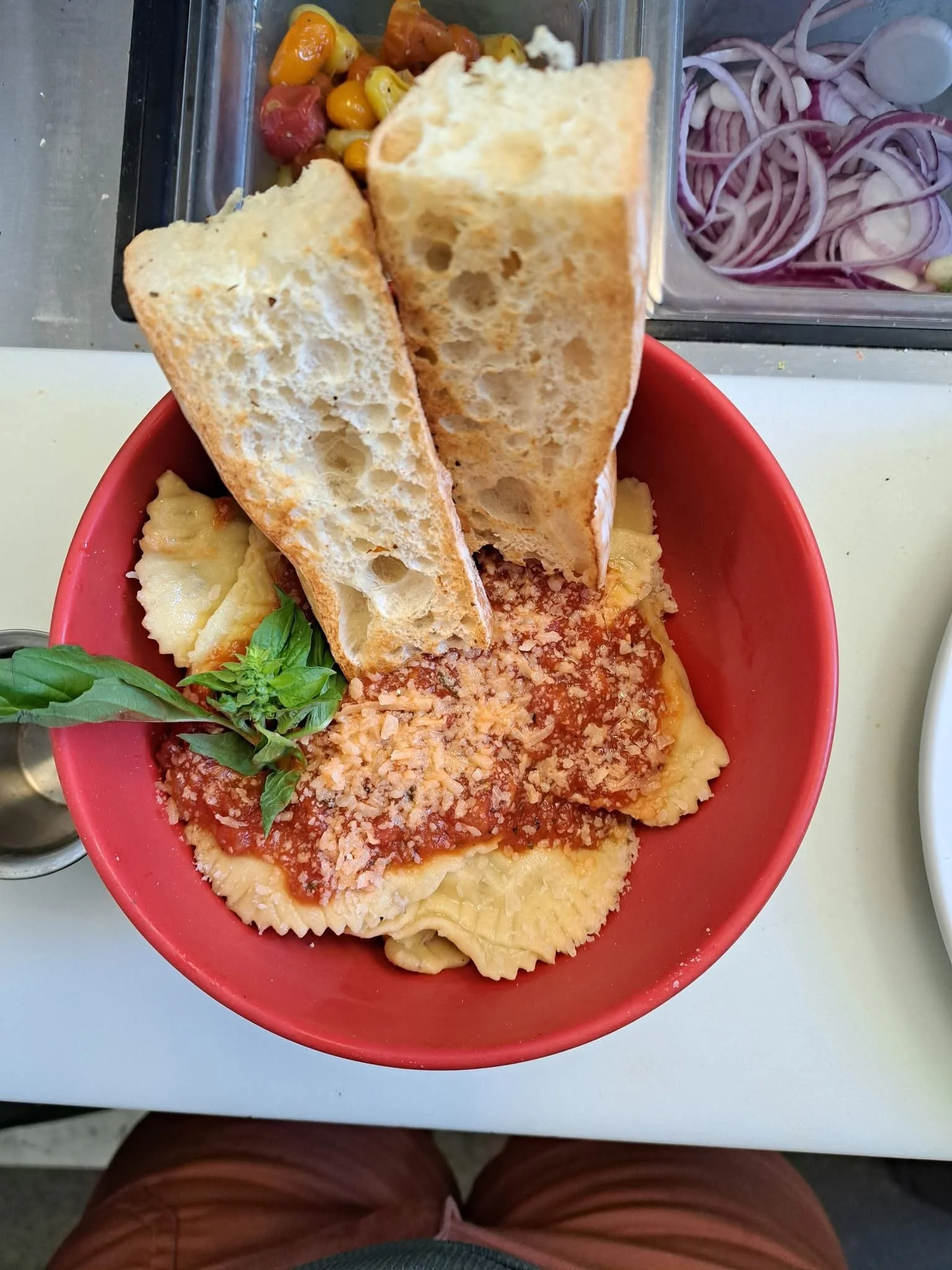 A red bowl containing cooked ravioli pasta topped with marinara sauce and grated cheese, garnished with basil, and two slices of toasted bread. In the background, there is a container of mixed roasted vegetables and a container of sliced onions.