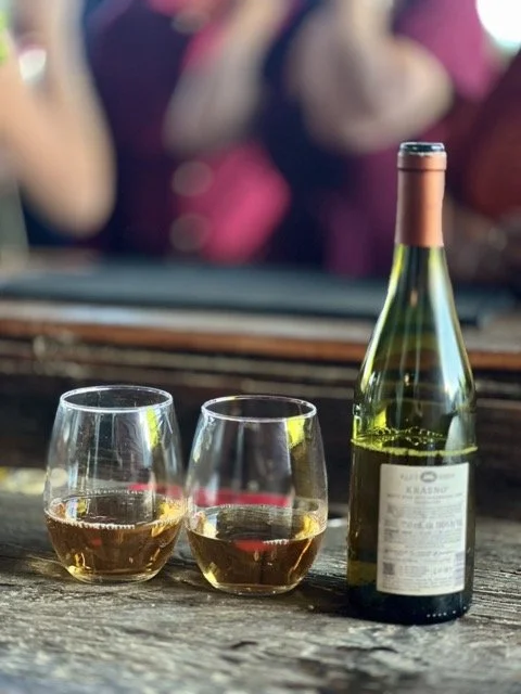 A partially filled wine bottle and two glasses of white wine on a rustic wooden surface, with a blurred background of people at a social gathering.