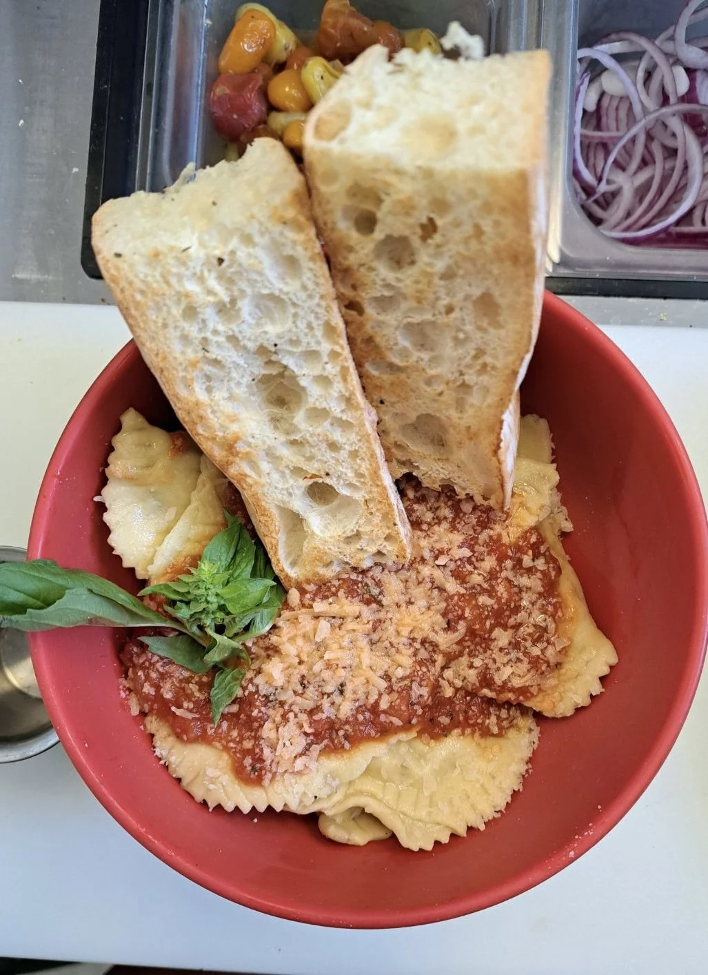 A red bowl containing cooked ravioli pasta topped with marinara sauce and grated cheese, garnished with basil, and two slices of toasted bread. In the background, there is a container of mixed roasted vegetables and a container of sliced onions.