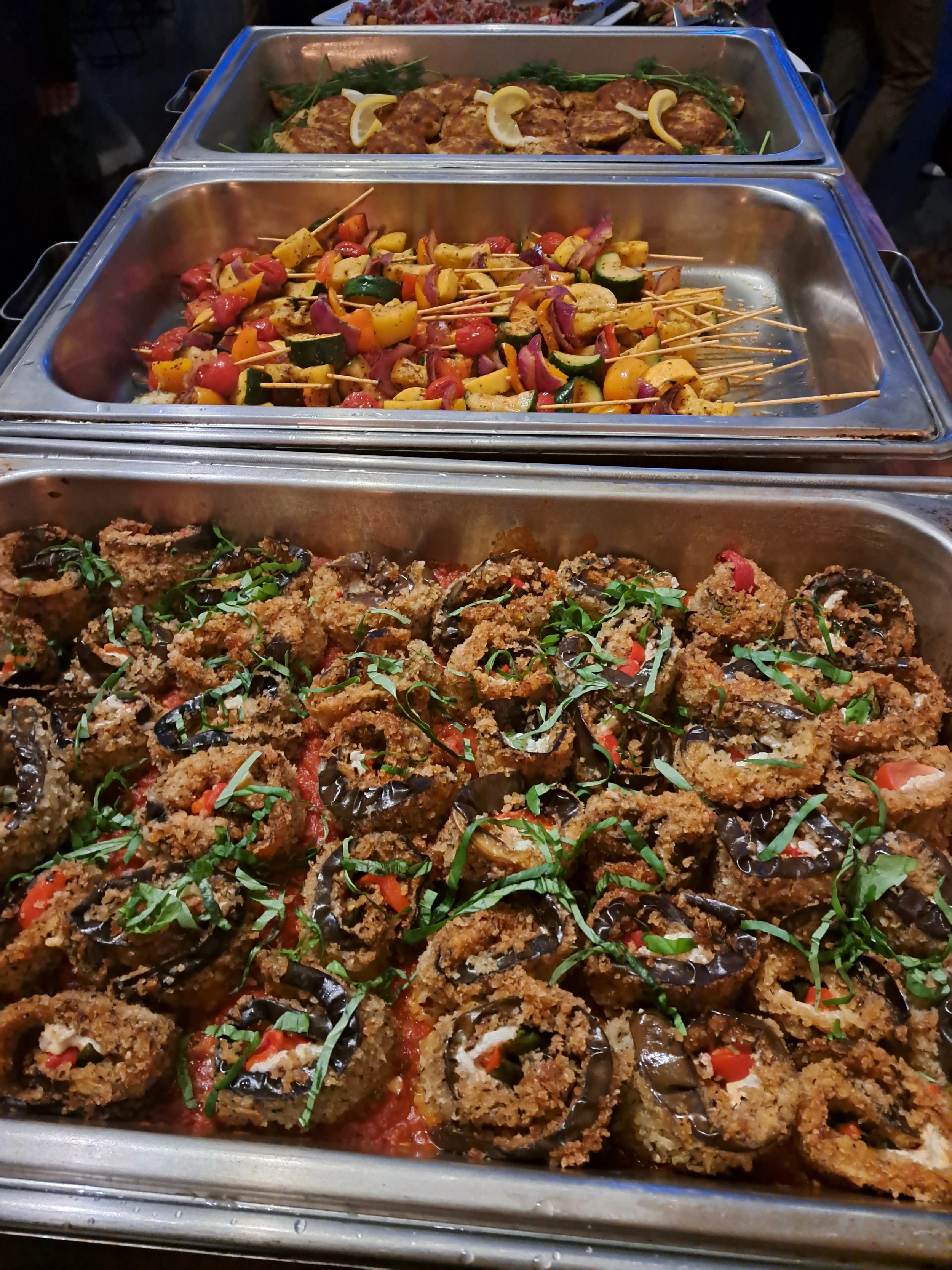 Buffet containing fried eggplant slices topped with herbs, skewers of grilled vegetables including zucchini, cherry tomatoes, bell peppers, and onions, and breaded chicken or fish fillets garnished with chopped herbs.