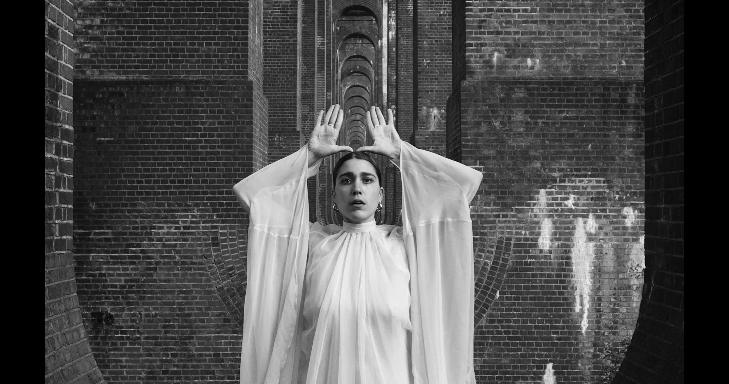 A woman dressed in a flowing, light-colored dress stands against a brick archway, holding her hands above her head with fingers spread apart, looking directly at the camera with a serious expression.