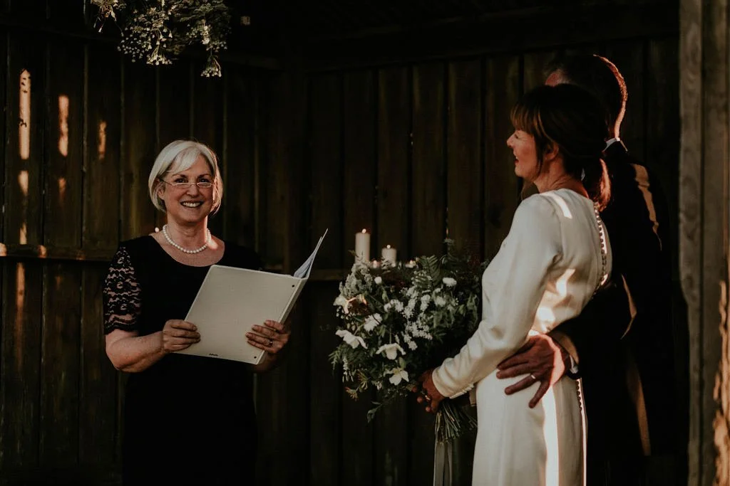 A wedding ceremony with a woman officiating, holding a folder and smiling, a couple is standing together with the bride holding a floral bouquet.