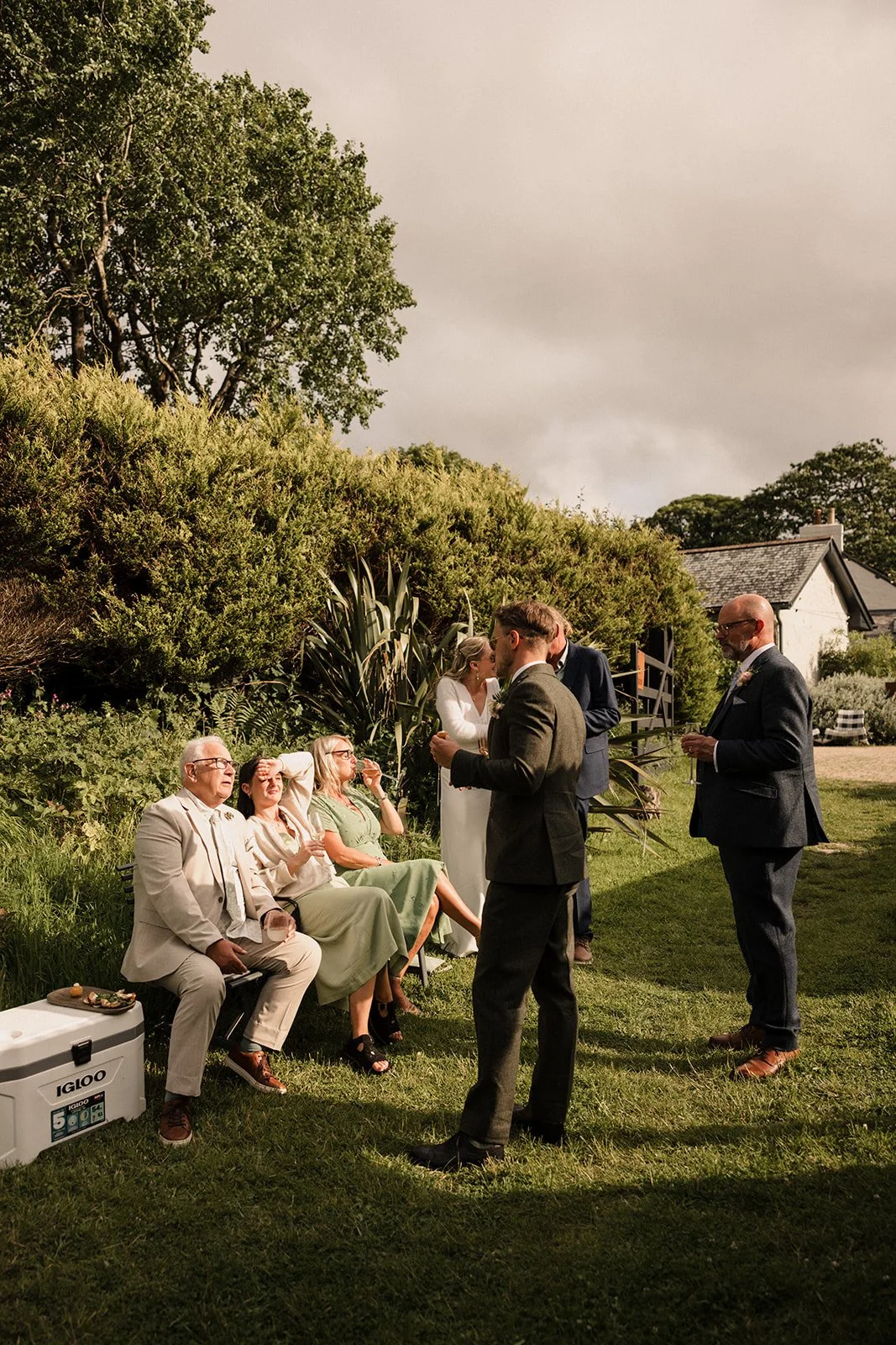 People at an outdoor wedding reception, some seated on a bench and others standing, with a lush green background and a cloudy sky.