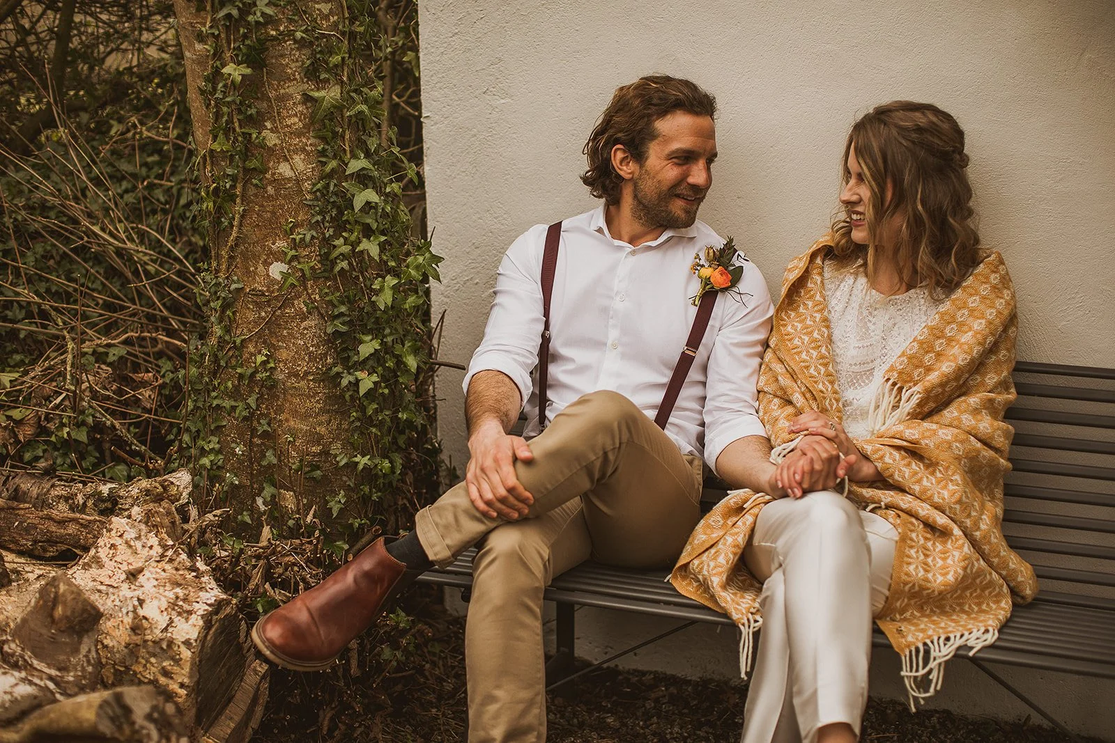 Couple sitting on a bench, man in white shirt and suspenders, woman wrapped in a patterned blanket, holding hands, outdoors with ivy-covered tree in background.
