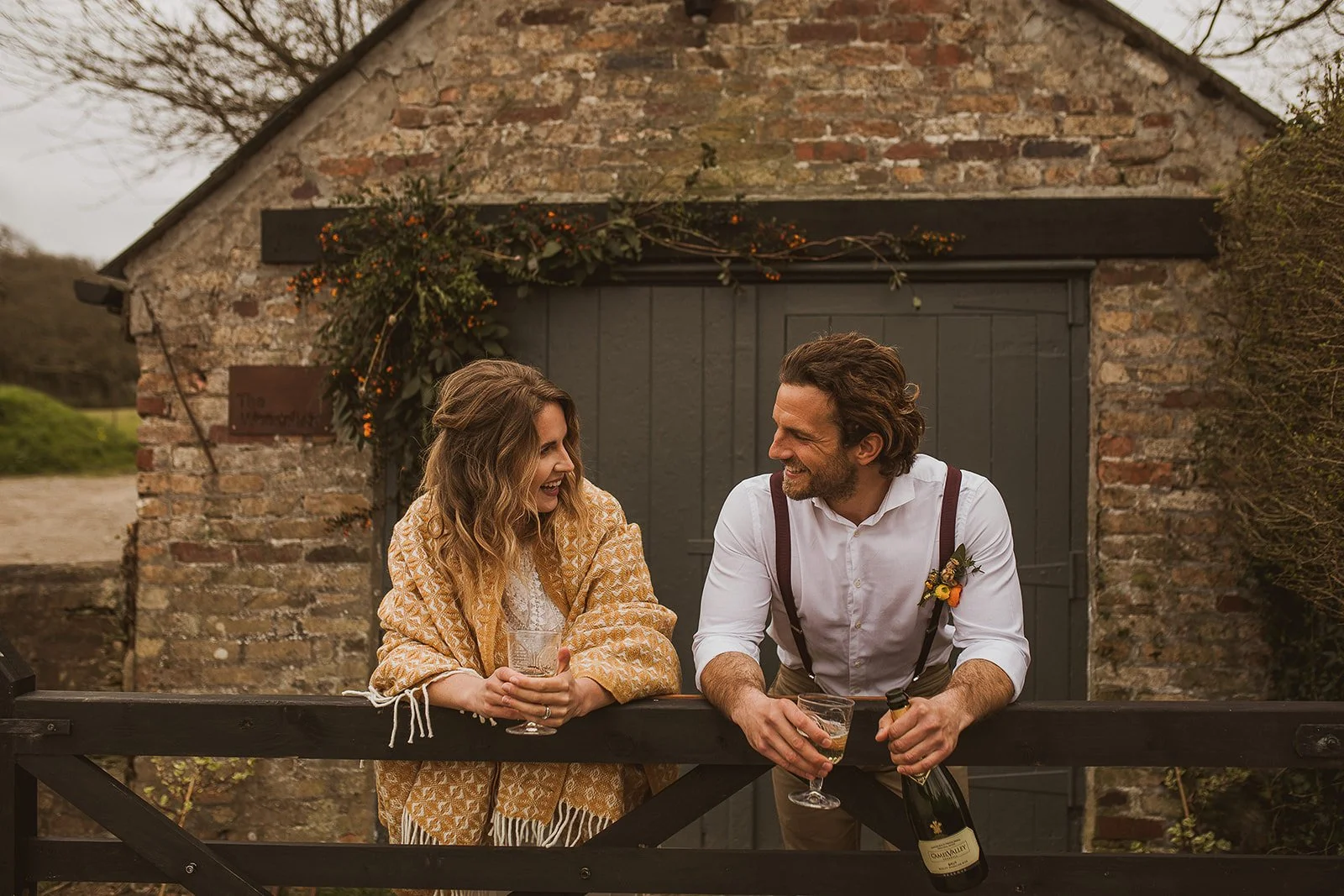 Couple smiling and leaning on a wooden fence, holding glasses of wine, with a rustic brick building in the background and vine decorations above the door.