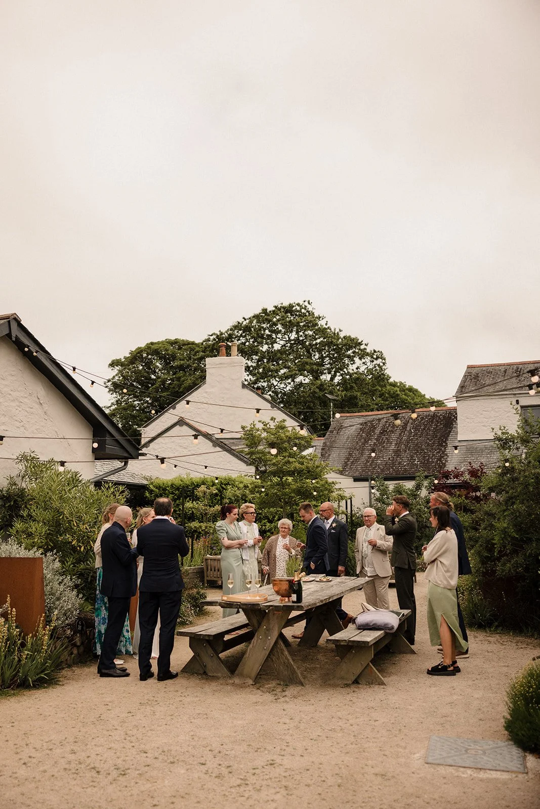 A group of people in formal attire gathered outdoors around a wooden table during a social event, with string lights overhead and houses in the background.