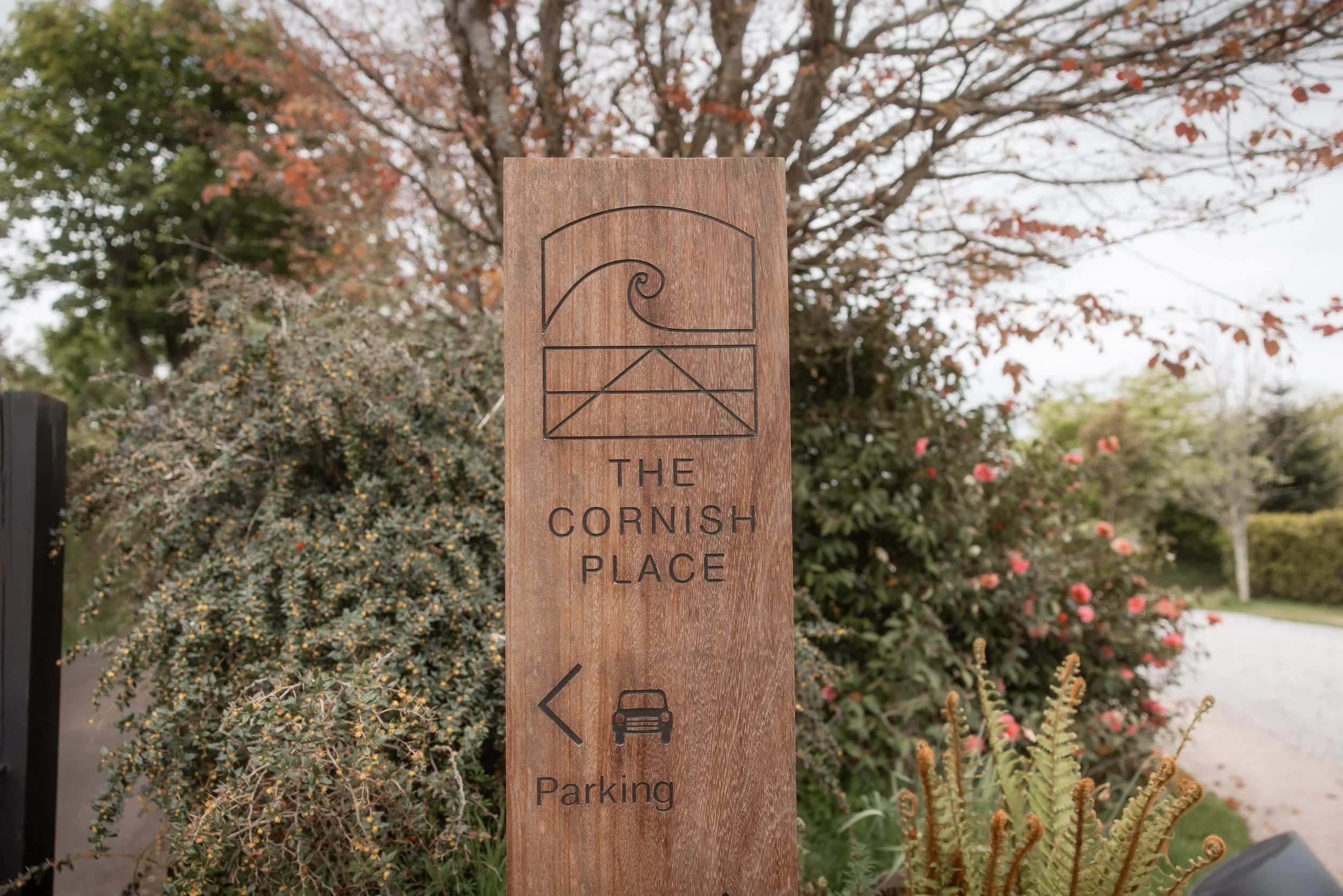 Wooden sign with a wave and envelope outline, text reading "The Cornish Place," and an arrow pointing left with a car icon and the word "Parking," set amidst greenery and flowering bushes.