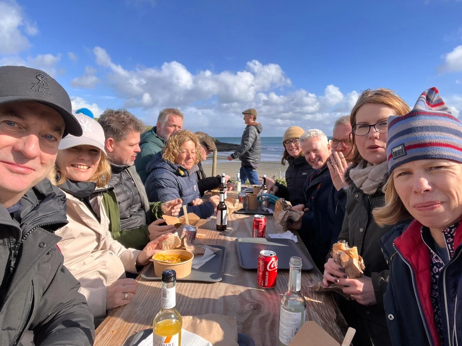 Group of people sitting at an outdoor table near the beach on a sunny day, eating and drinking.
