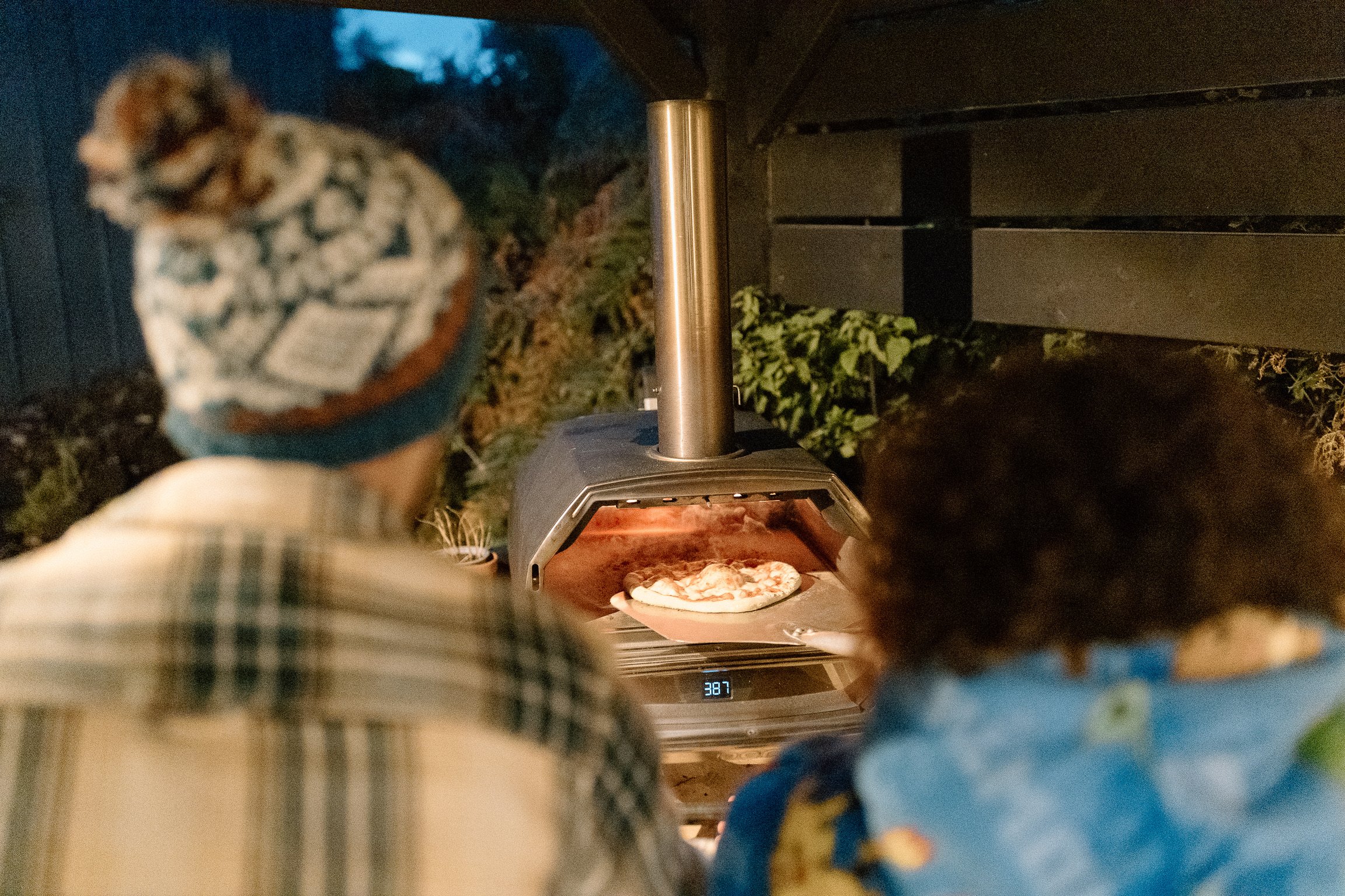 Two children watching pizza bake in an outdoor pizza oven during evening, with one child wearing a hat and plaid shirt, and the other with curly hair and a patterned shirt.