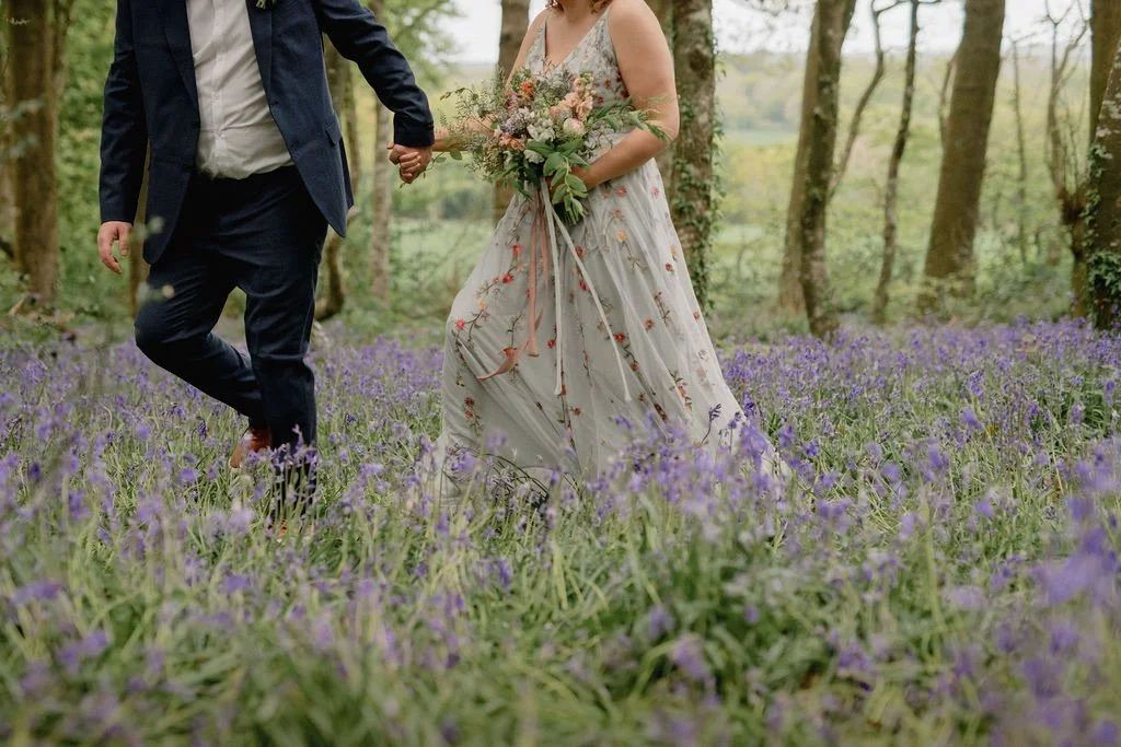 Couple holding hands walking through a forest meadow with purple flowers, woman in a floral dress holding a bouquet.