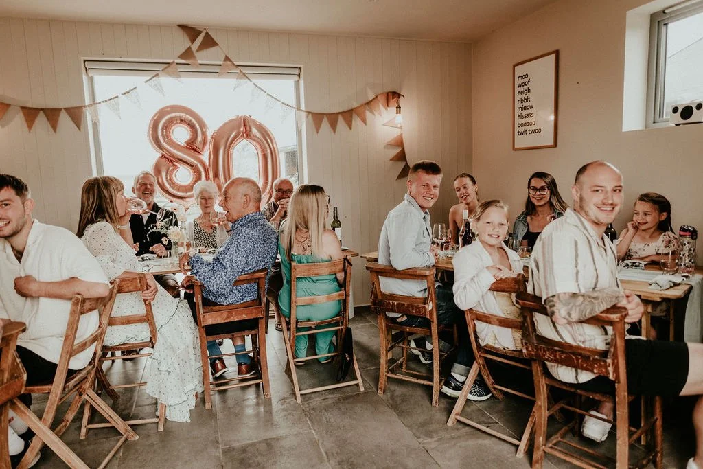 A birthday celebration with around 12 people sitting at two wooden tables in a cozy room. The decor includes large rose gold balloons spelling out "80" and a banner with triangular flags. The group appears cheerful, some with drinks, and there is a f