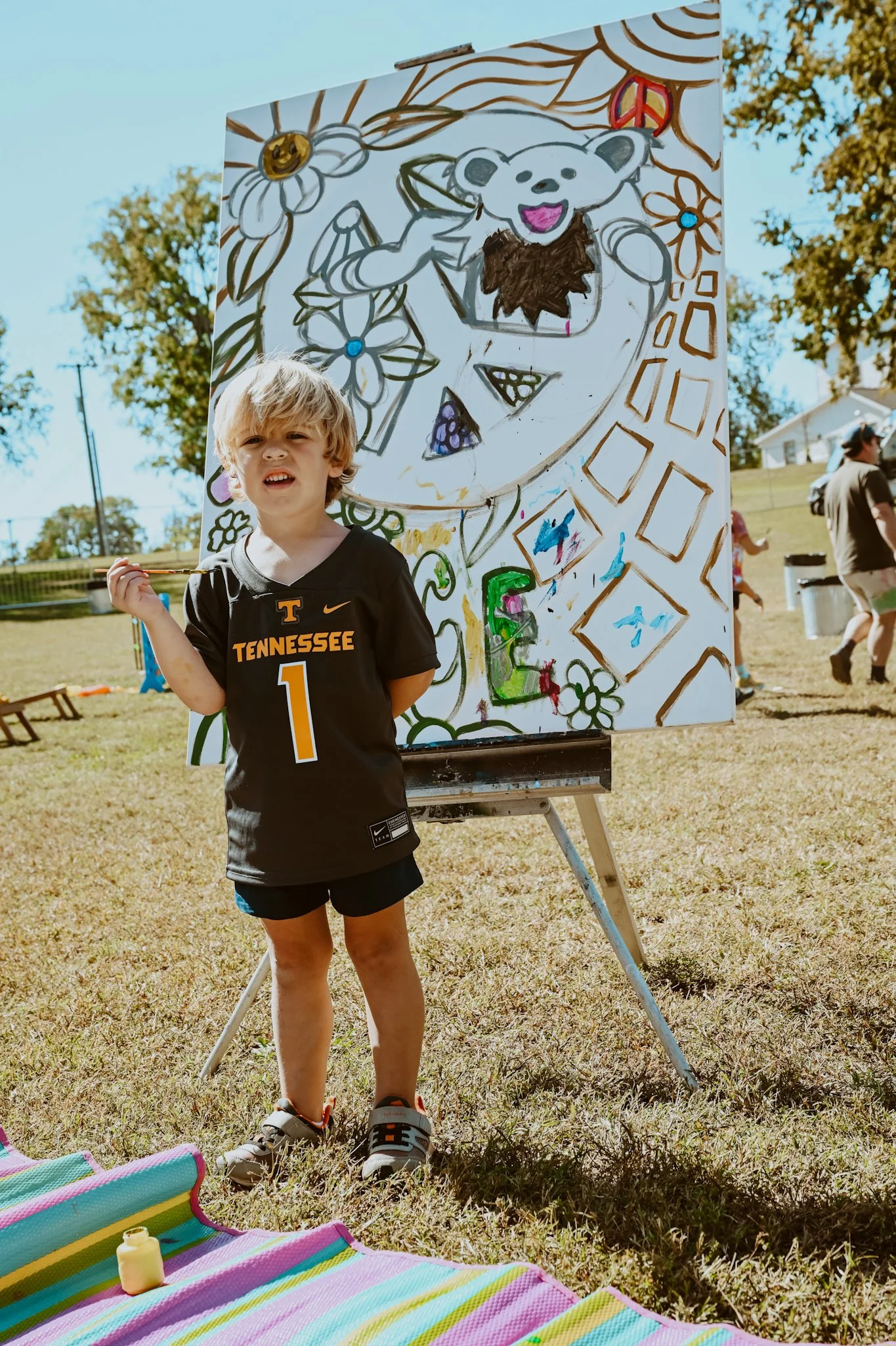 A young boy wearing a Tennessee Volunteers football jersey stands next to a large outdoor painting on an easel, depicting a cartoon bear, flowers, and abstract shapes, with a colorful striped blanket on the ground in front of him.