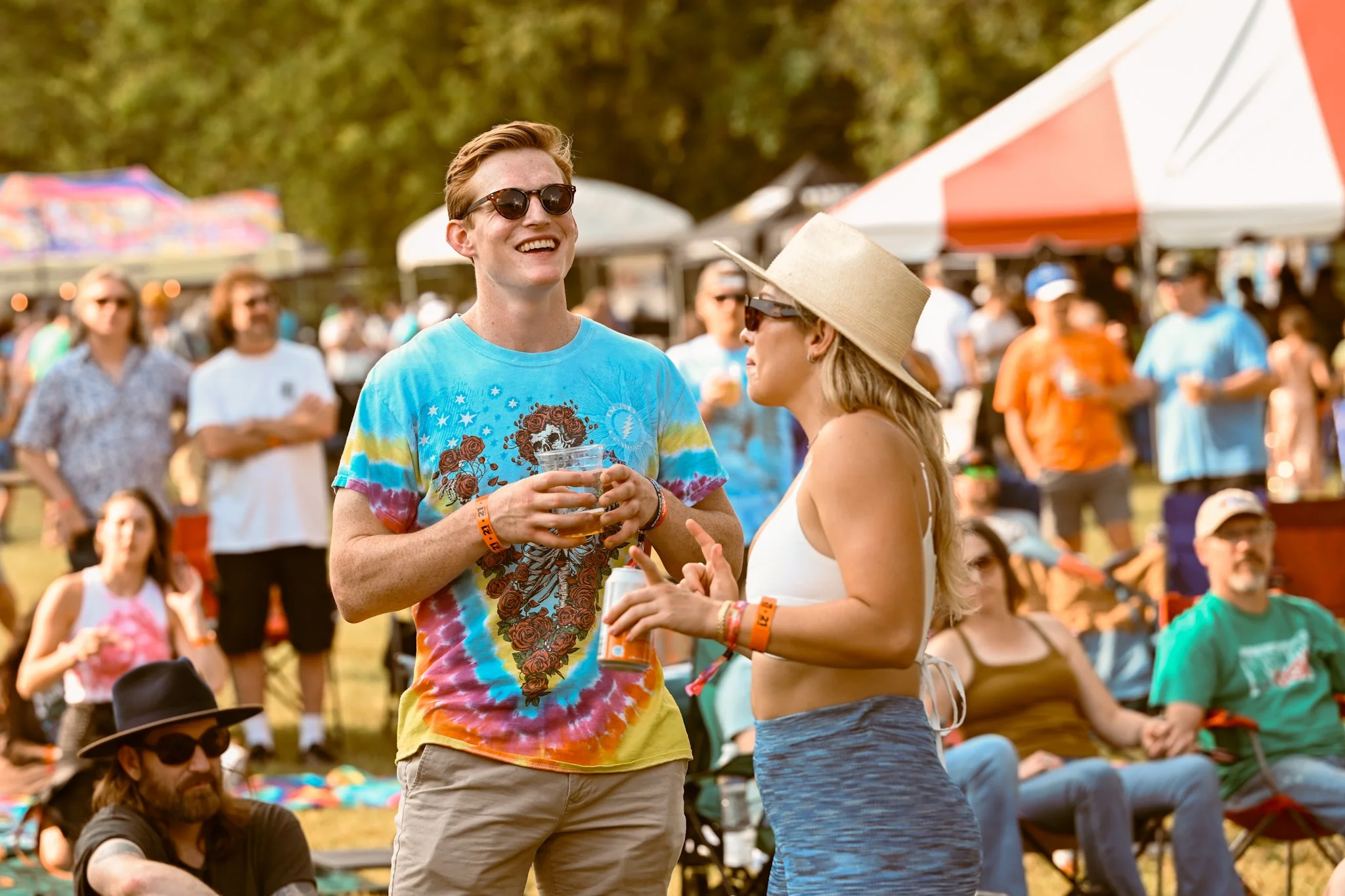 Two young adults, a man and a woman, are talking and smiling at an outdoor festival or concert. The man is wearing a colorful tie-dye t-shirt with a skull design, sunglasses, and has short red hair. The woman is wearing a large straw hat, sunglasses,