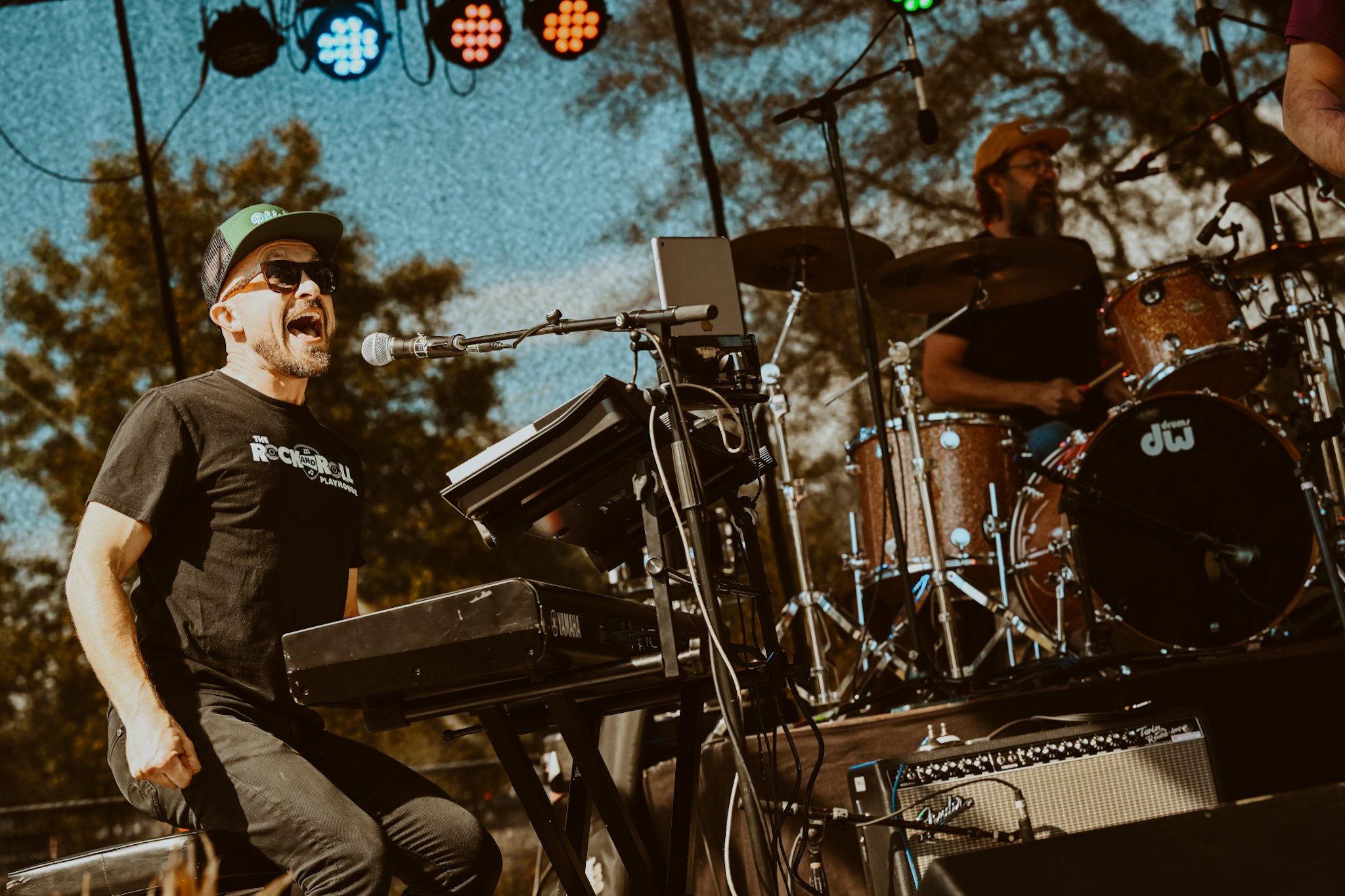 Musicians performing on outdoor stage, one playing keyboard and singing into a microphone, another on drums in background, trees and blue sky in background.