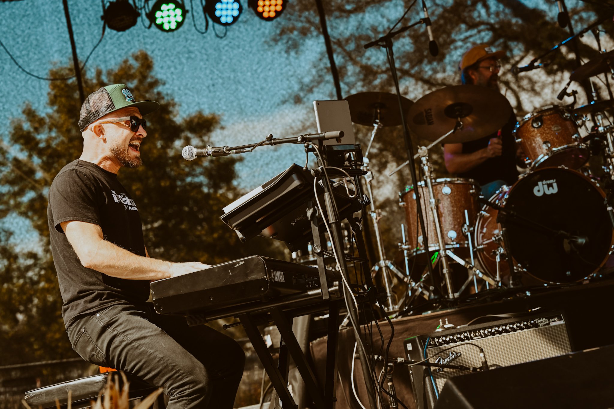 Musicians performing outdoors on stage, one playing keyboard and singing into a microphone, the other playing drums in the background, with stage lights and trees visible.