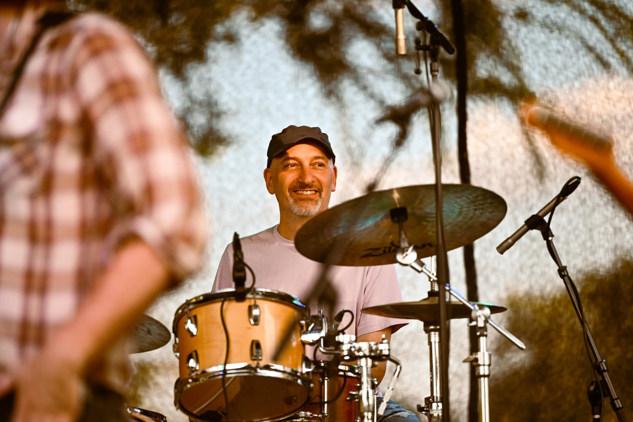 A man smiling while sitting behind a drum set on a stage, with audience members in the foreground.