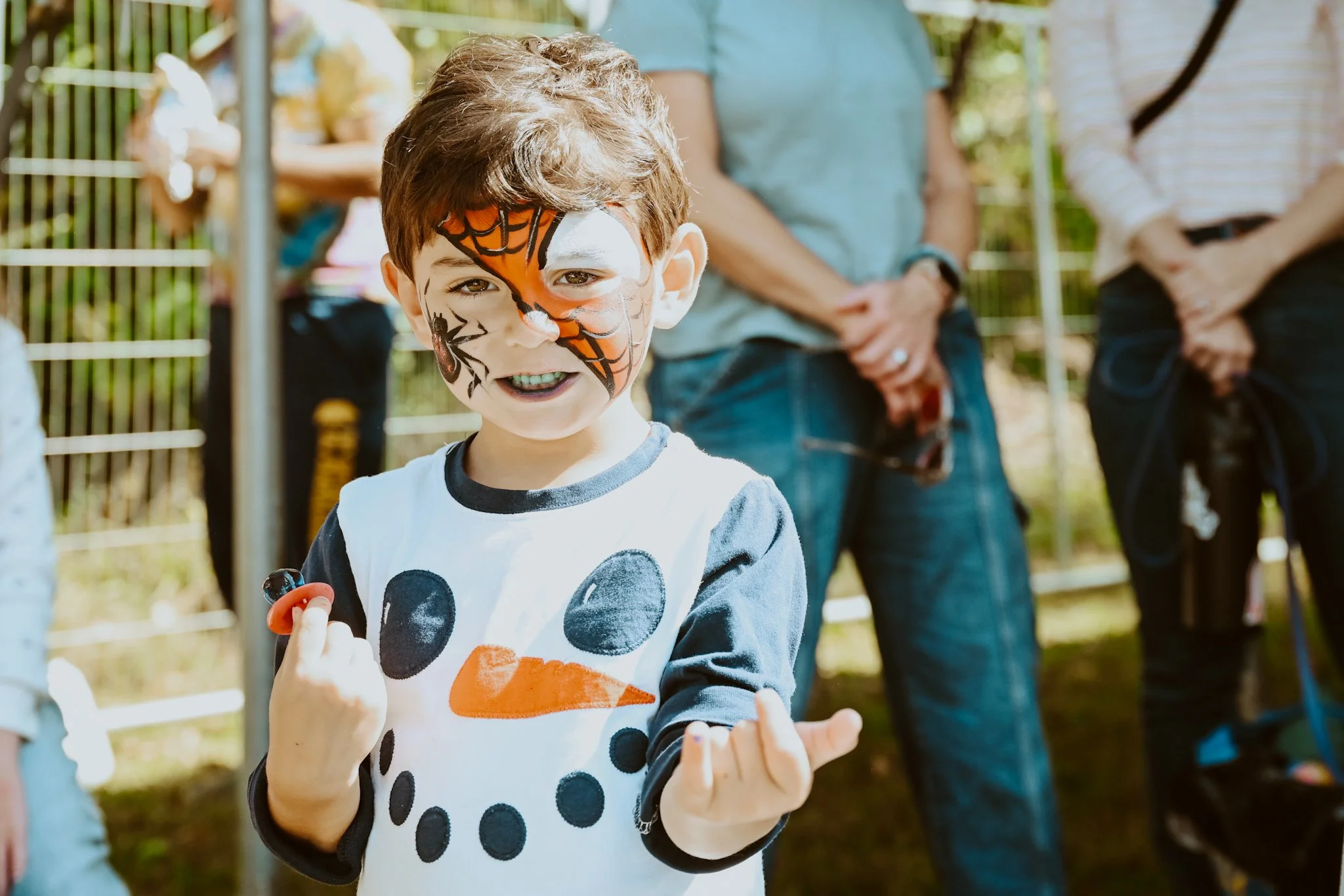 A young boy with face paint resembling Spider-Man's mask and a snowman shirt, smiling and holding a marker, outside during daytime with adults in the background.