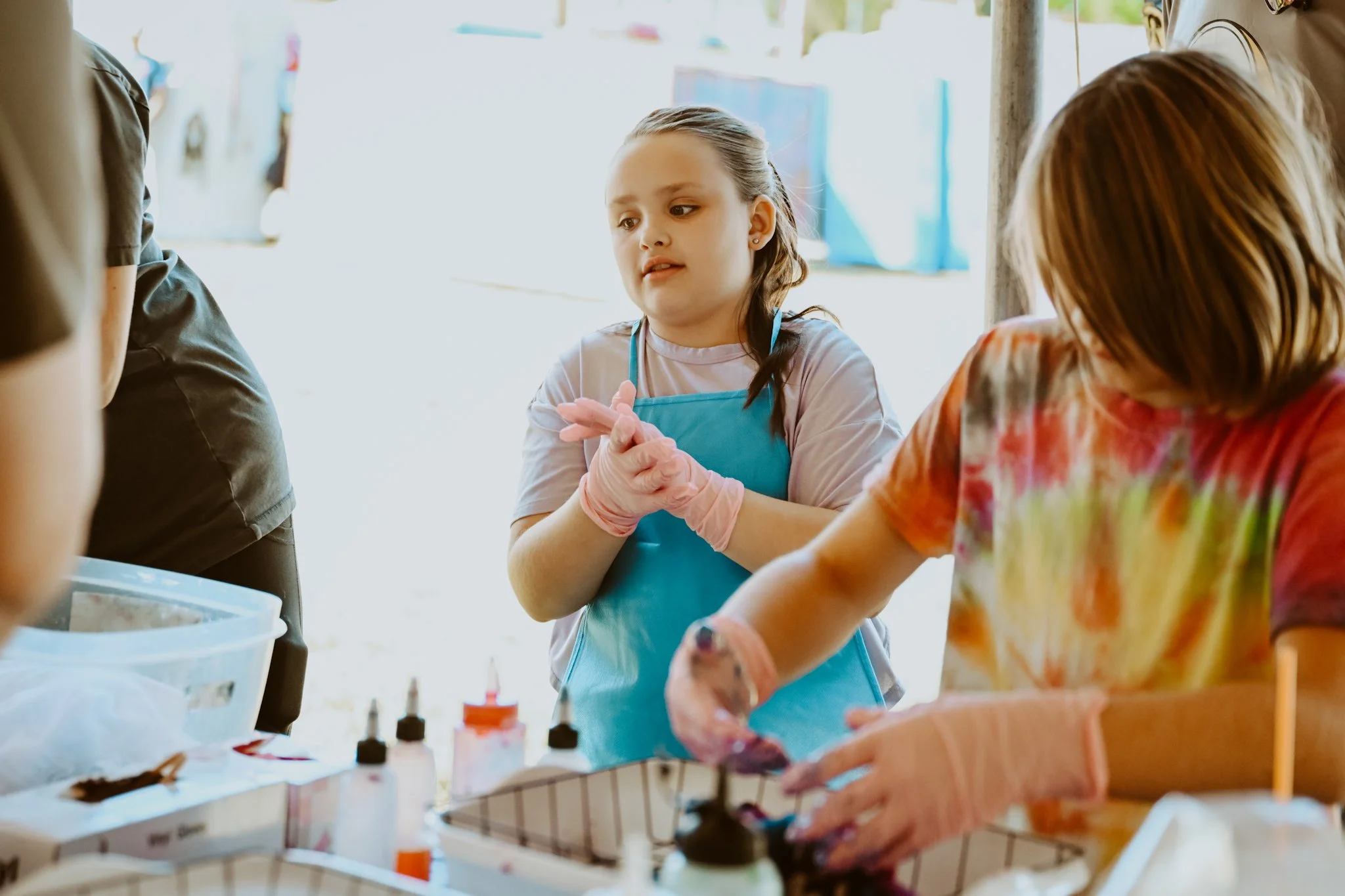 Two young girls covered in colorful tie-dye and wearing gloves are participating in a tie-dye activity at a table with bottles of dye and other supplies.