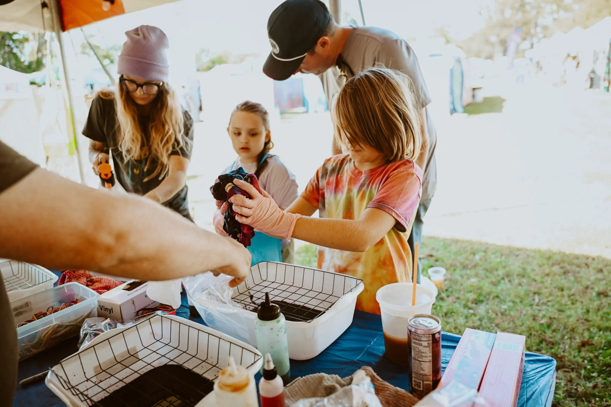 Group of children and adults gathered around a table making tie-dye shirts outdoors.