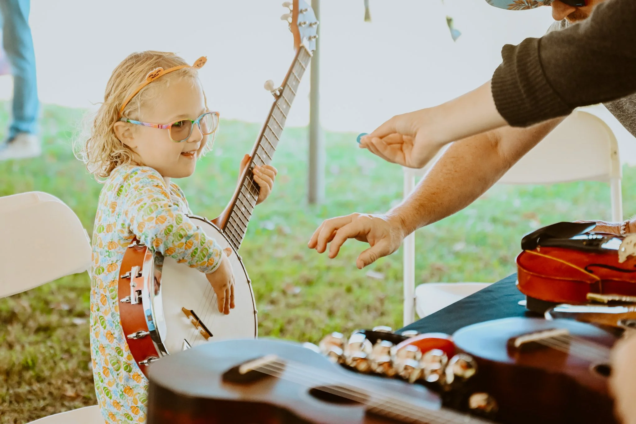A young girl wearing sunglasses and a floral dress receives a blue coin from an adult at an outdoor music event. She is holding a guitar-shaped instrument and is smiling. There are other musical instruments and a table in front of them.