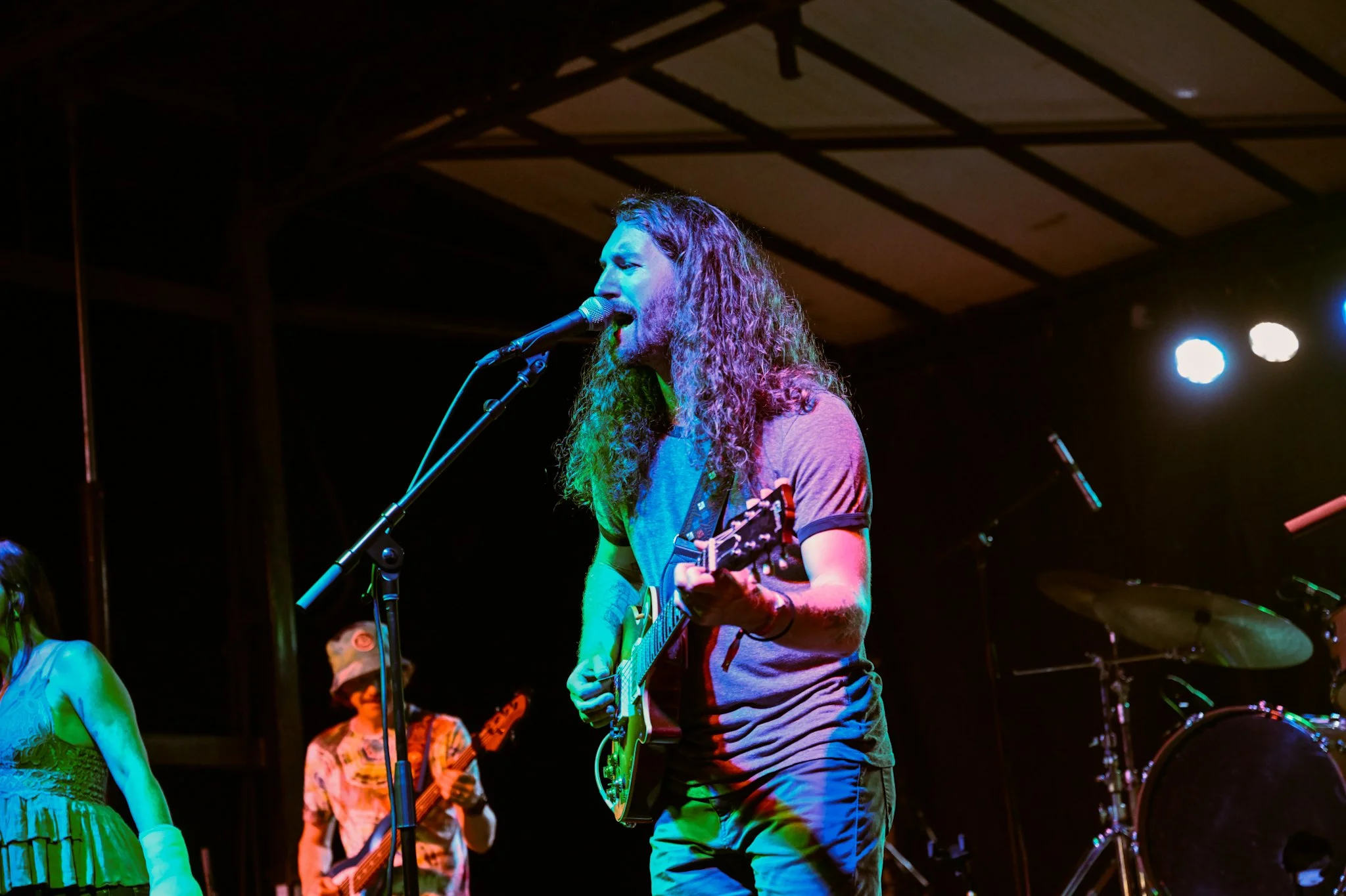 A male musician with long curly hair singing into a microphone while playing an electric guitar on stage. Two other musicians are partially visible in the background, both playing guitars. The stage is lit with colorful lights, and a drum set is visi
