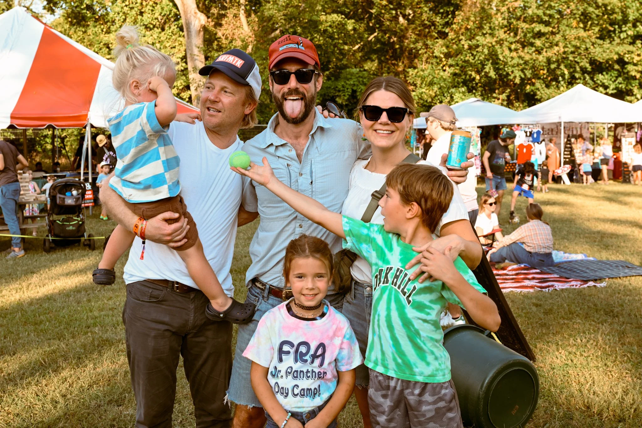 A group of happy people, including children and adults, at an outdoor festival or fair with tents and trees in the background.