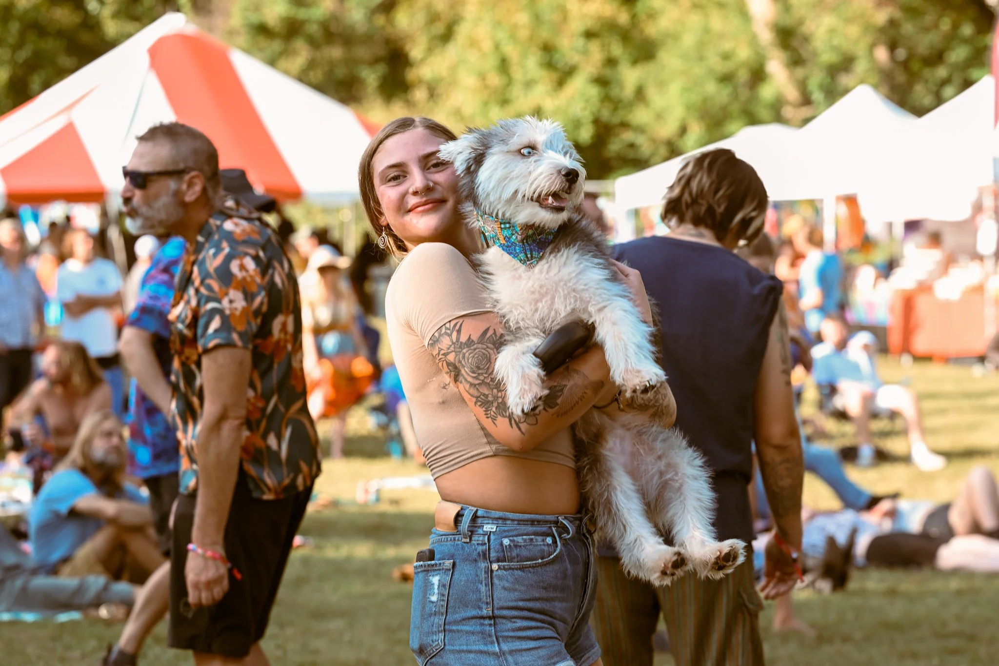 A woman with tattoos holding a fluffy dog at an outdoor festival, with people, tents, and trees in the background.