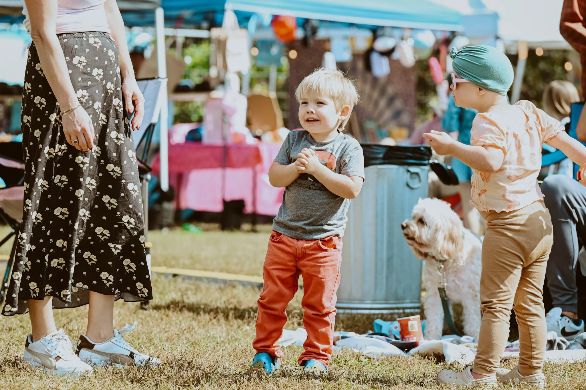 Two young children and a woman at an outdoor market, with tents and merchandise in the background. One child is smiling and clasping hands, while the other, wearing a green hat and sunglasses, is reaching out. A dog is nearby.