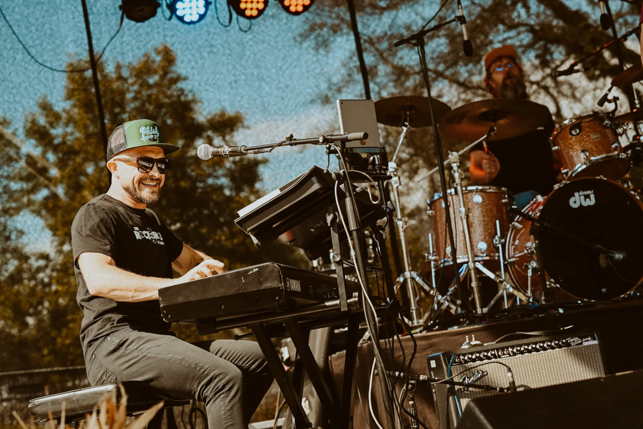 A man playing a keyboard and a drummer performing on a stage outdoors with trees in the background during daytime.