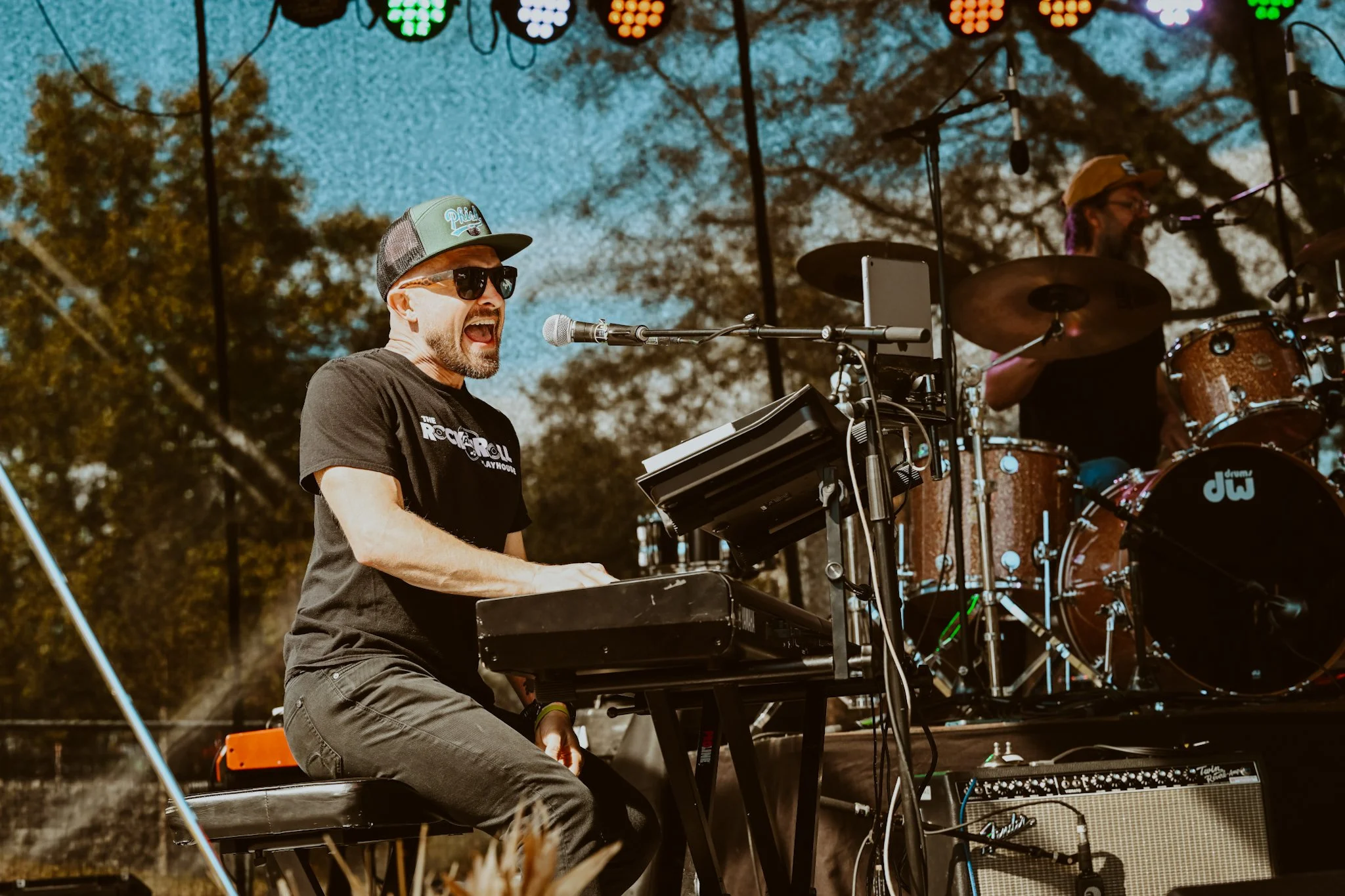 A musician wearing sunglasses and a cap, passionately singing into a microphone while playing a keyboard during an outdoor concert in daytime. A drummer is visible in the background playing drums.