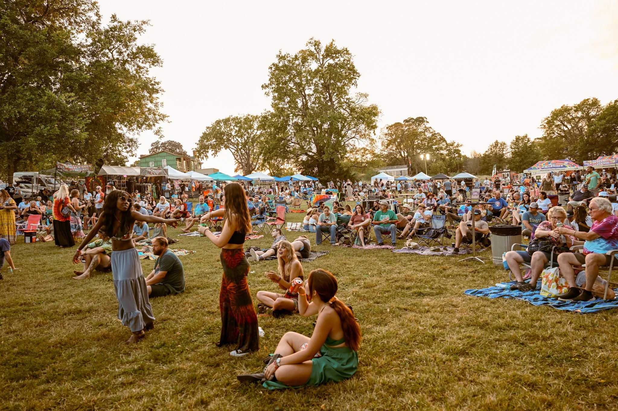 Crowd attending outdoor festival or event in a park during sunset, with people sitting on grass, dancing, and socializing near tents and food stands.
