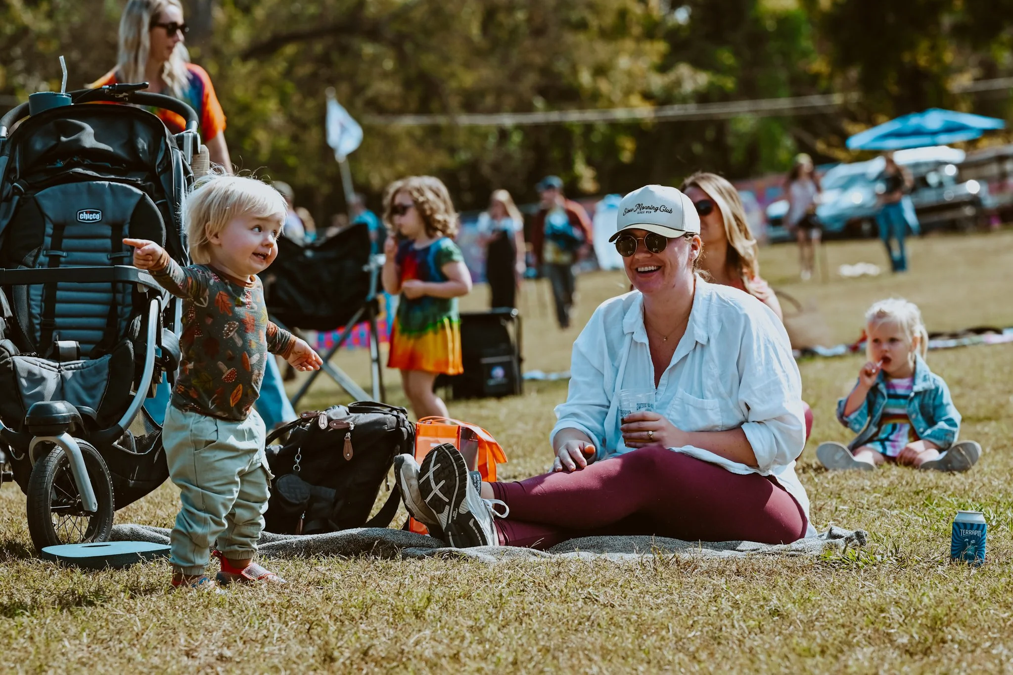 A woman sitting on a blanket smiling as she interacts with a young child standing nearby in a park during daytime. Other children and adults are in the background, with tents and cars visible.