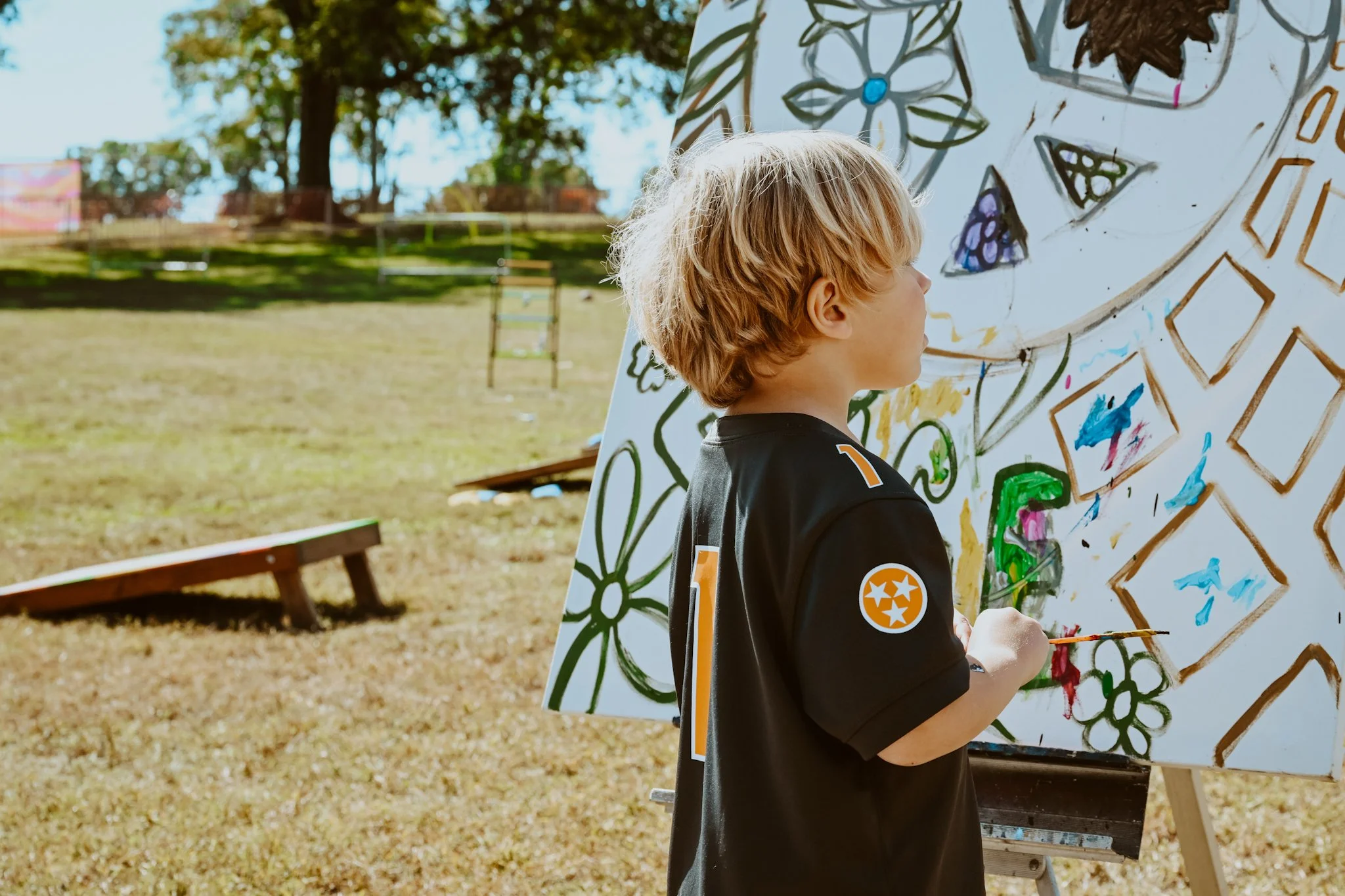 A young boy with blonde hair painting on a large outdoor canvas featuring colorful floral and geometric designs during daytime in a park.