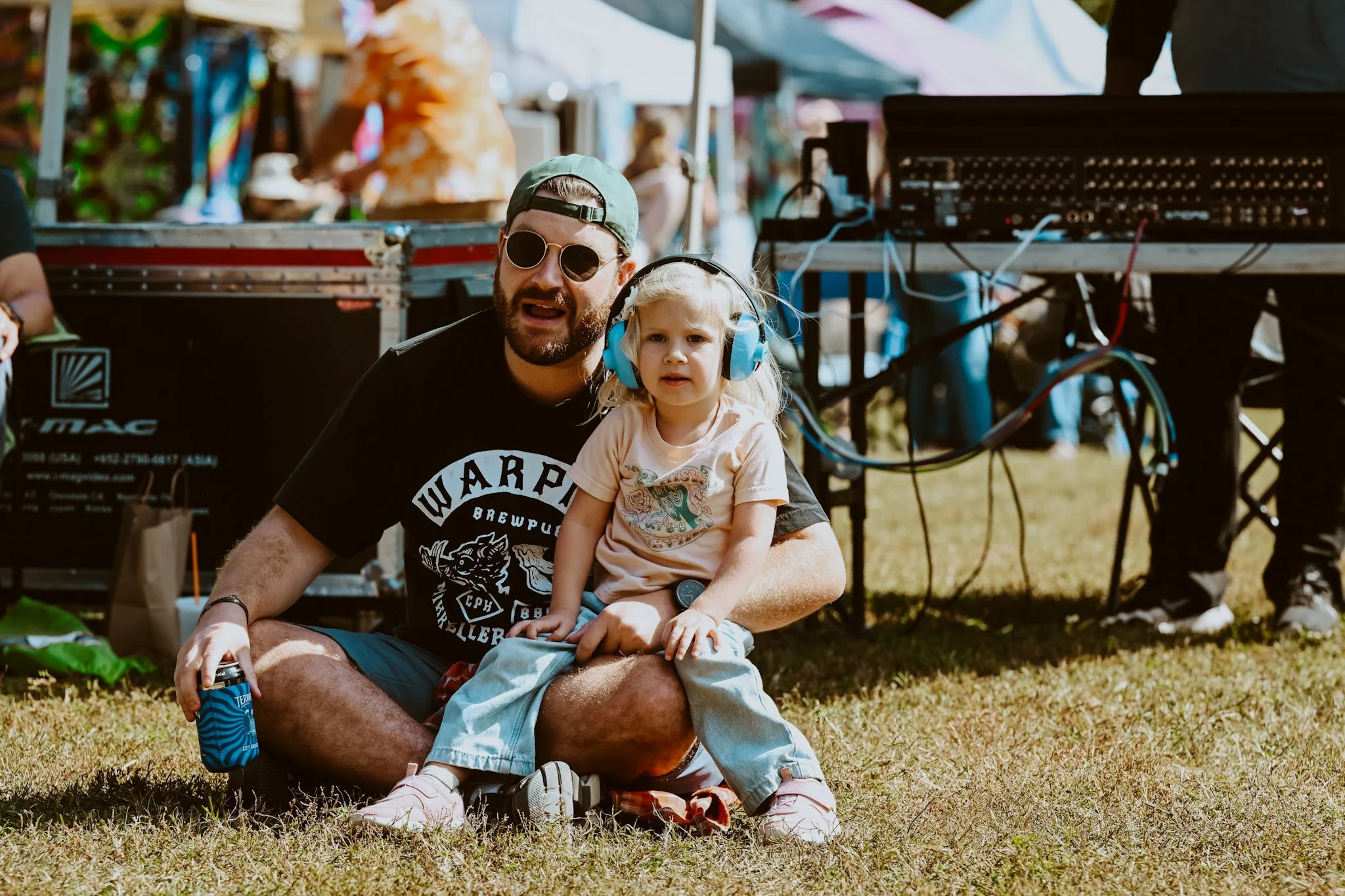 A man with a beard, wearing sunglasses, a green cap, a black t-shirt, and shorts, sitting on the grass. He is holding a young girl with blonde hair, wearing large blue ear protection, a light pink t-shirt with a dinosaur graphic, and light pants. The