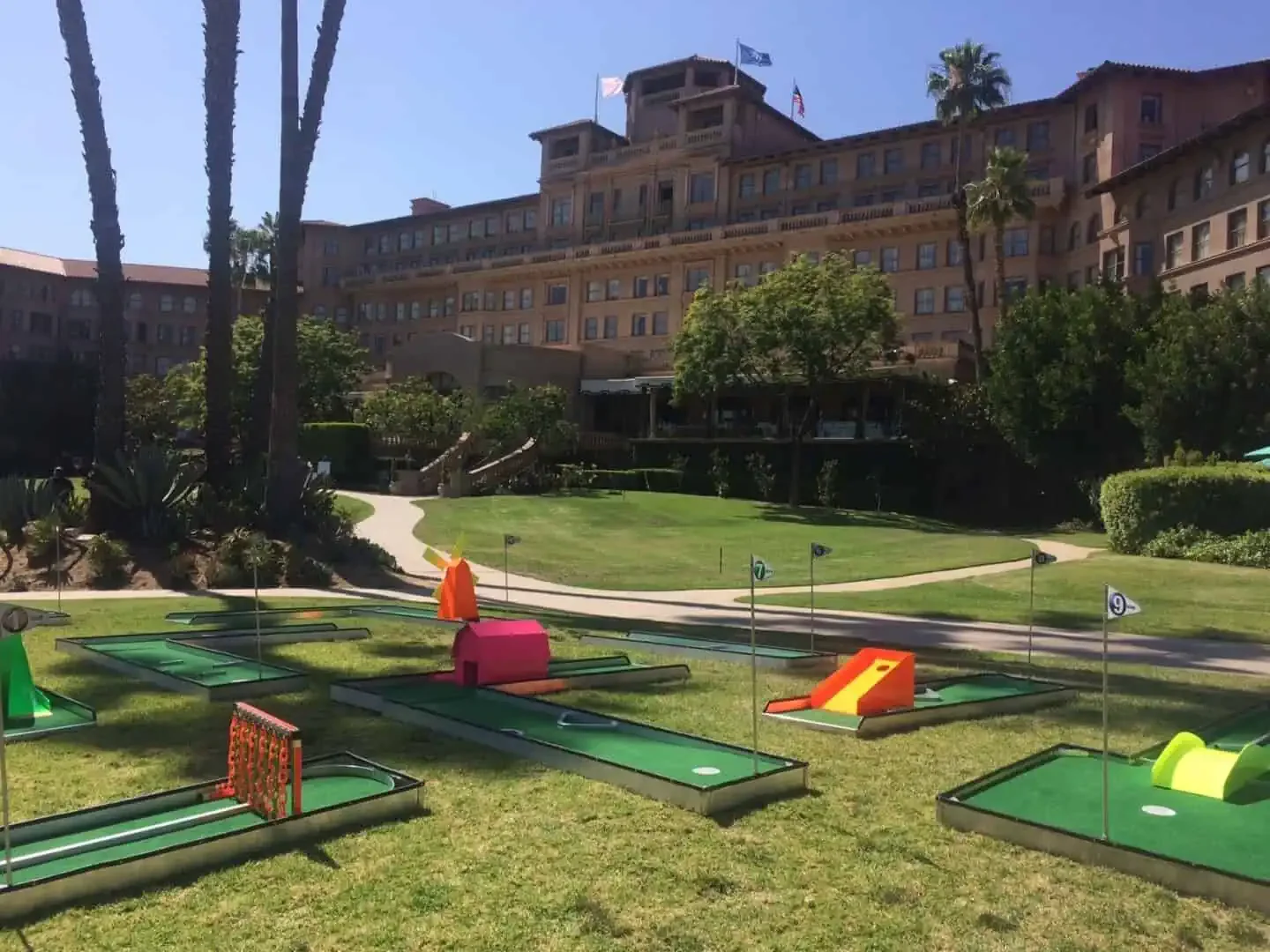 A young boy with blond hair climbing a rock wall outdoors, wearing a harness and smiling at the camera, with tents and a table in the background on a sunny day.