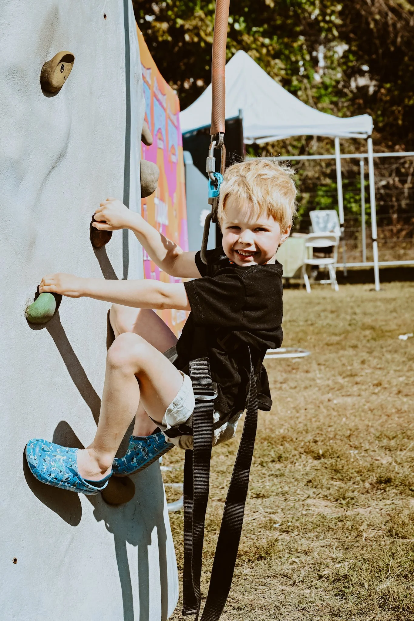 Young boy climbing an outdoor rock wall, smiling, with safety harness in a park or outdoor event setting.
