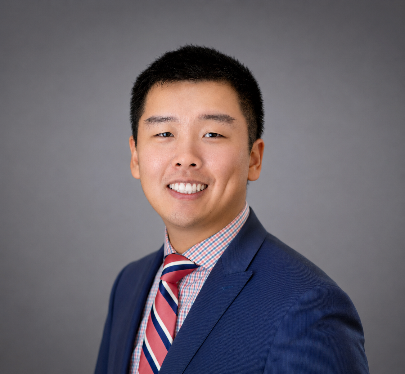 Professional headshot of a young Asian man with short black hair, wearing a dark blue suit, a checkered shirt, and a red, white, and blue striped tie, smiling against a gray background.