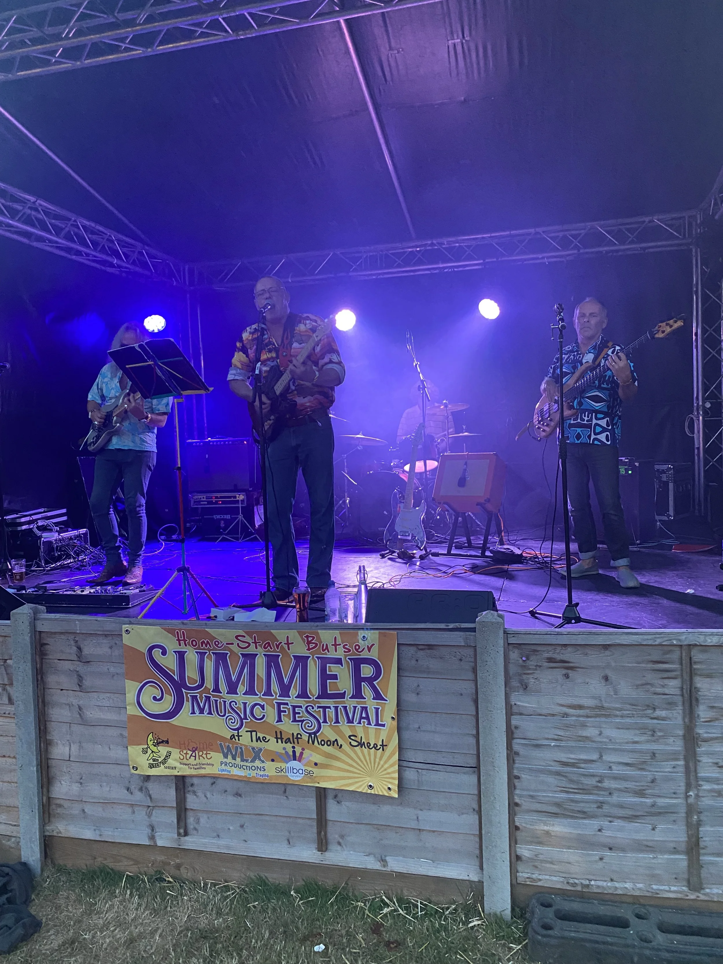 A band performing on stage at the Summer Music Festival, with four members playing instruments and singing, under purple and blue stage lights. A banner on the front fence reads 'Home-Start Butser Summer Music Festival.'