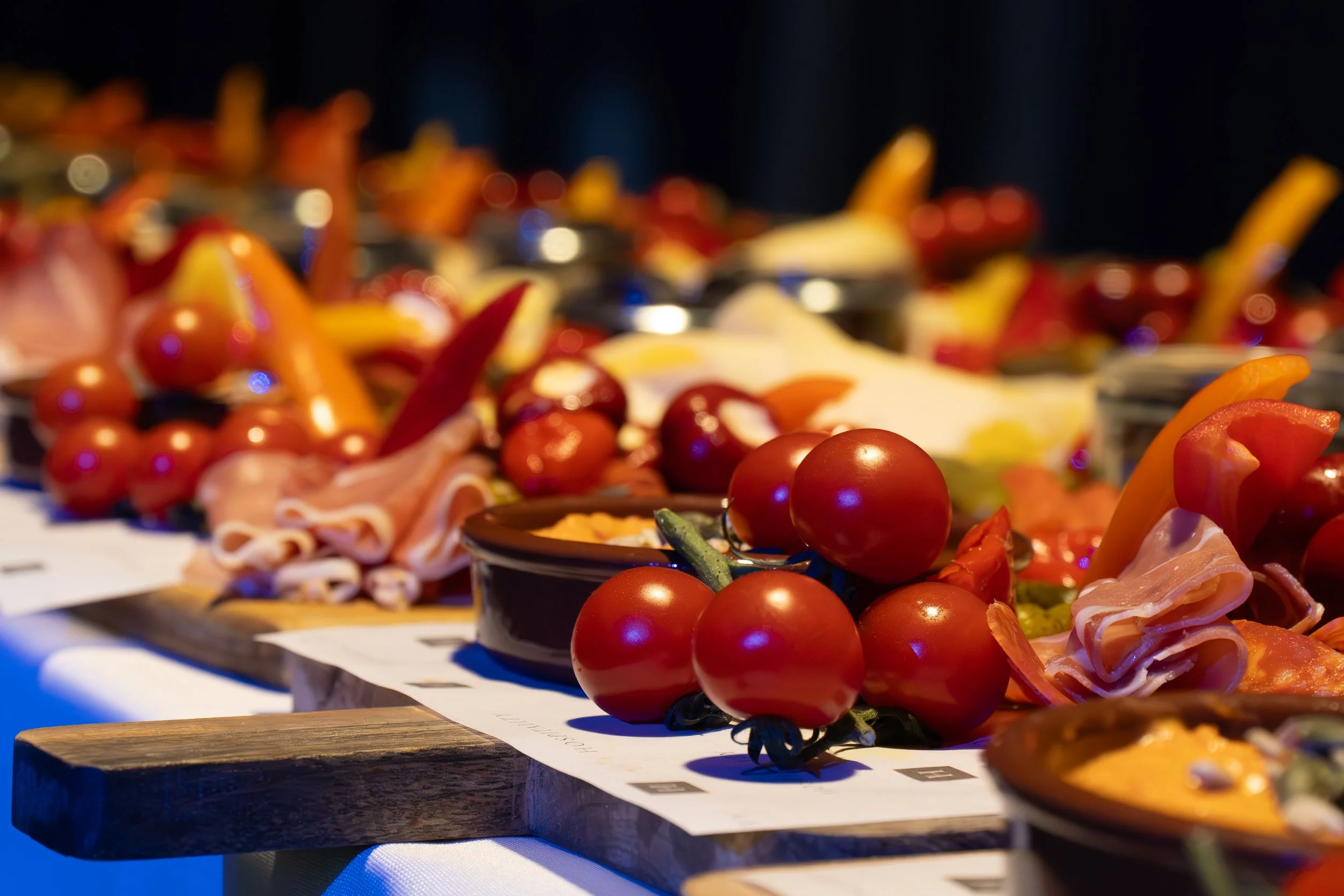 A display of fresh cherry tomatoes, sliced bell peppers, and cured meats on wooden boards at a food event.