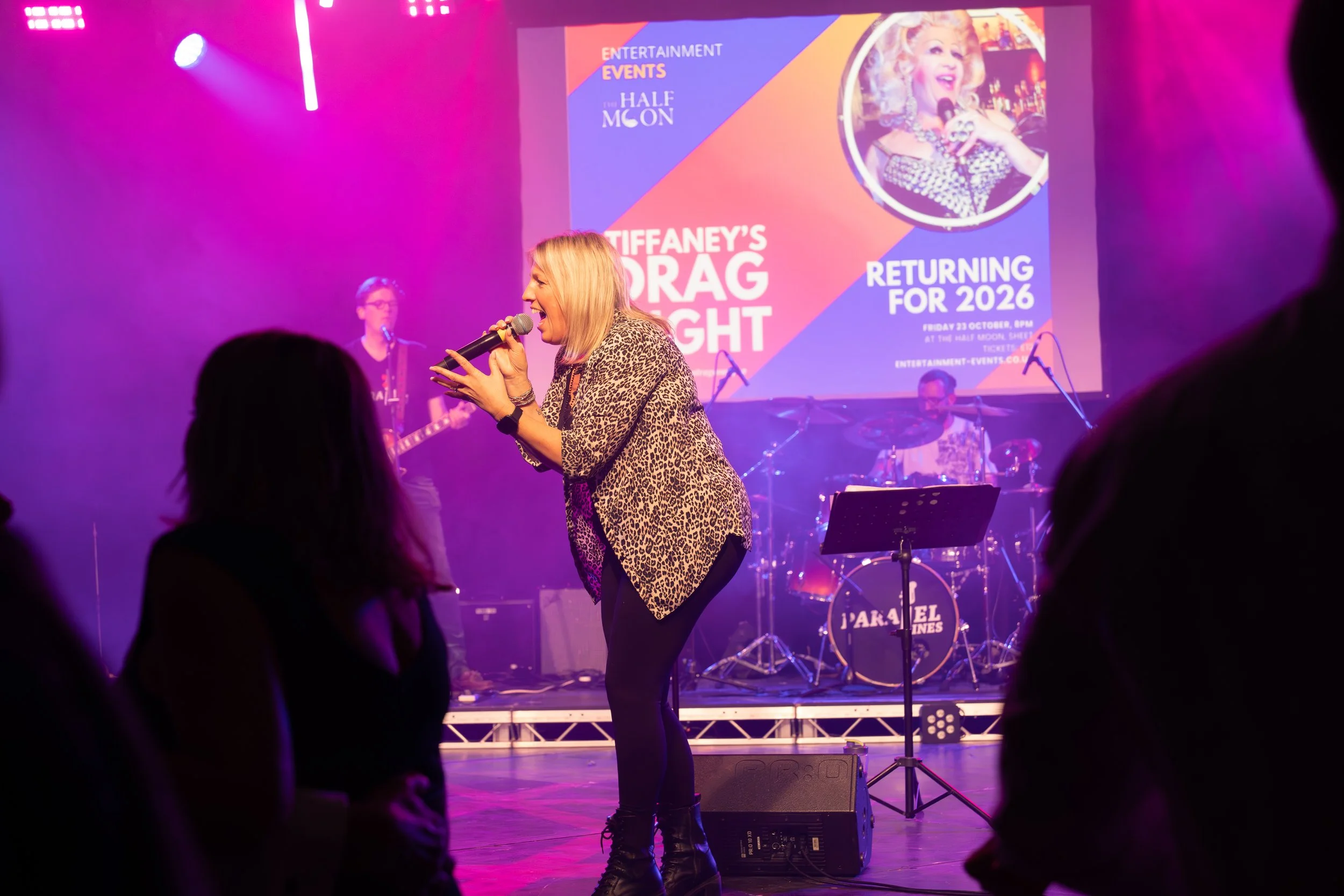 A woman singing on stage at a drag event, dressed in a leopard print jacket with a microphone. Behind her, a band plays with a guitarist, bassist, and drummer, and a large screen displays event information and a photo of another performer.